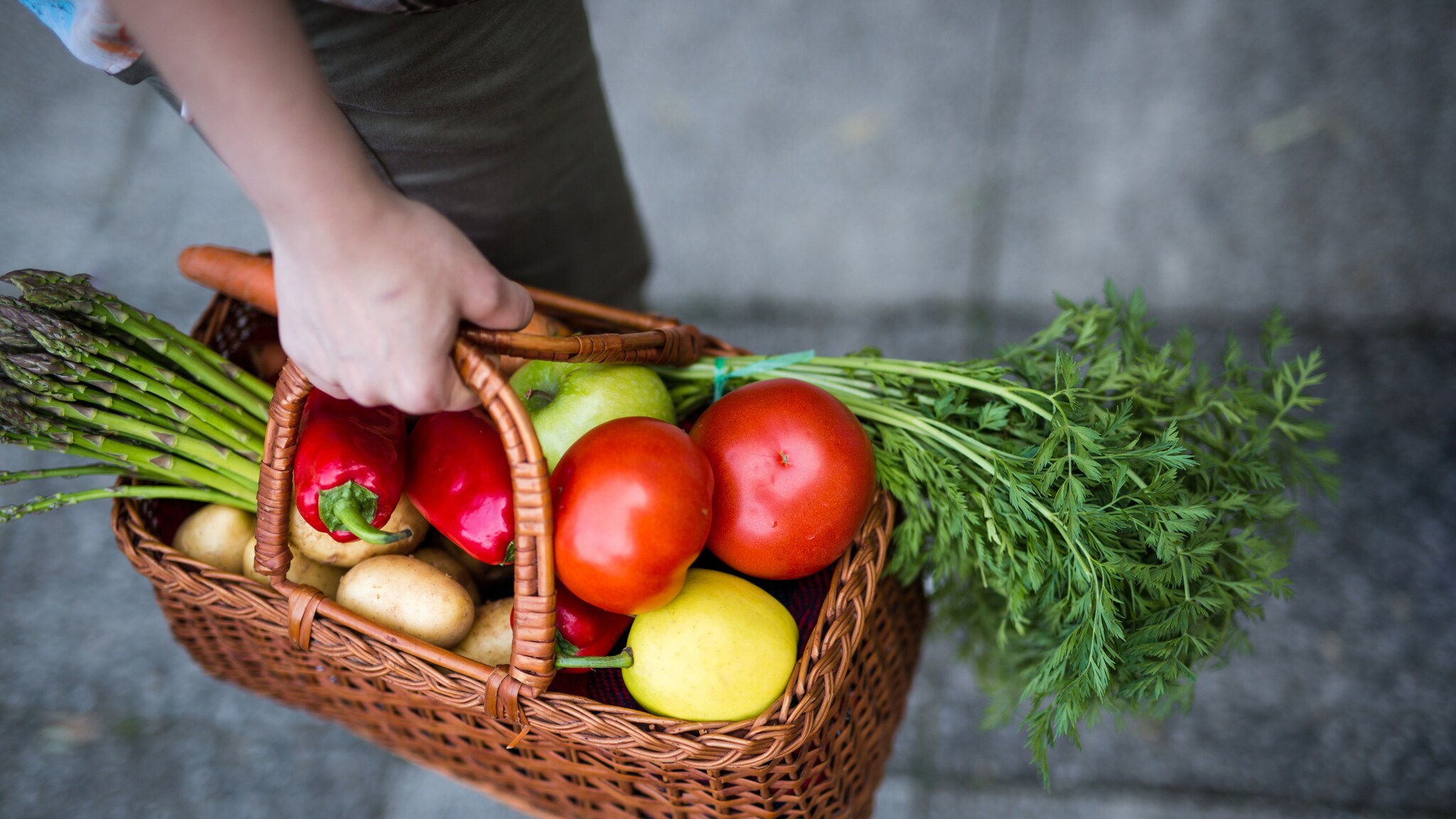 Hand hält geflochtenen Korb mit Biogemüse und Obst, darunter Tomaten, Karotten, Spargel, Paprika, Kartoffeln und ein Apfel.