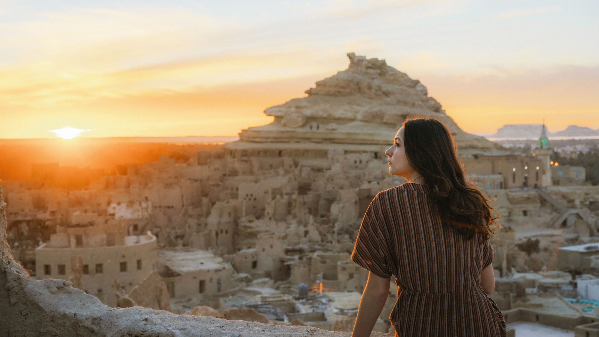 Frau mit braunem gestreiftem Kleid sitzt auf einer Mauer und blickt auf die Felsenstadt in der Siwa-Oase bei Sonnenuntergang in Ägypten. Frau mit braunem gestreiftem Kleid sitzt auf einer Mauer und blickt auf die Felsenstadt in der Siwa-Oase bei Sonnenuntergang in Ägypten.
