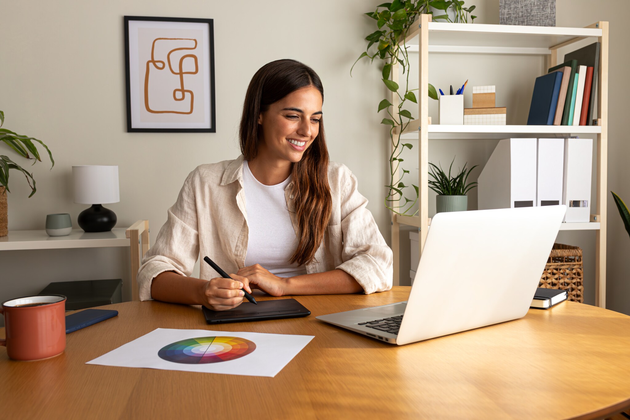 Eine lächelnde Frau arbeitet mit einem Grafiktablett an einem Laptop an einem Schreibtisch in einem Arbeitszimmer.