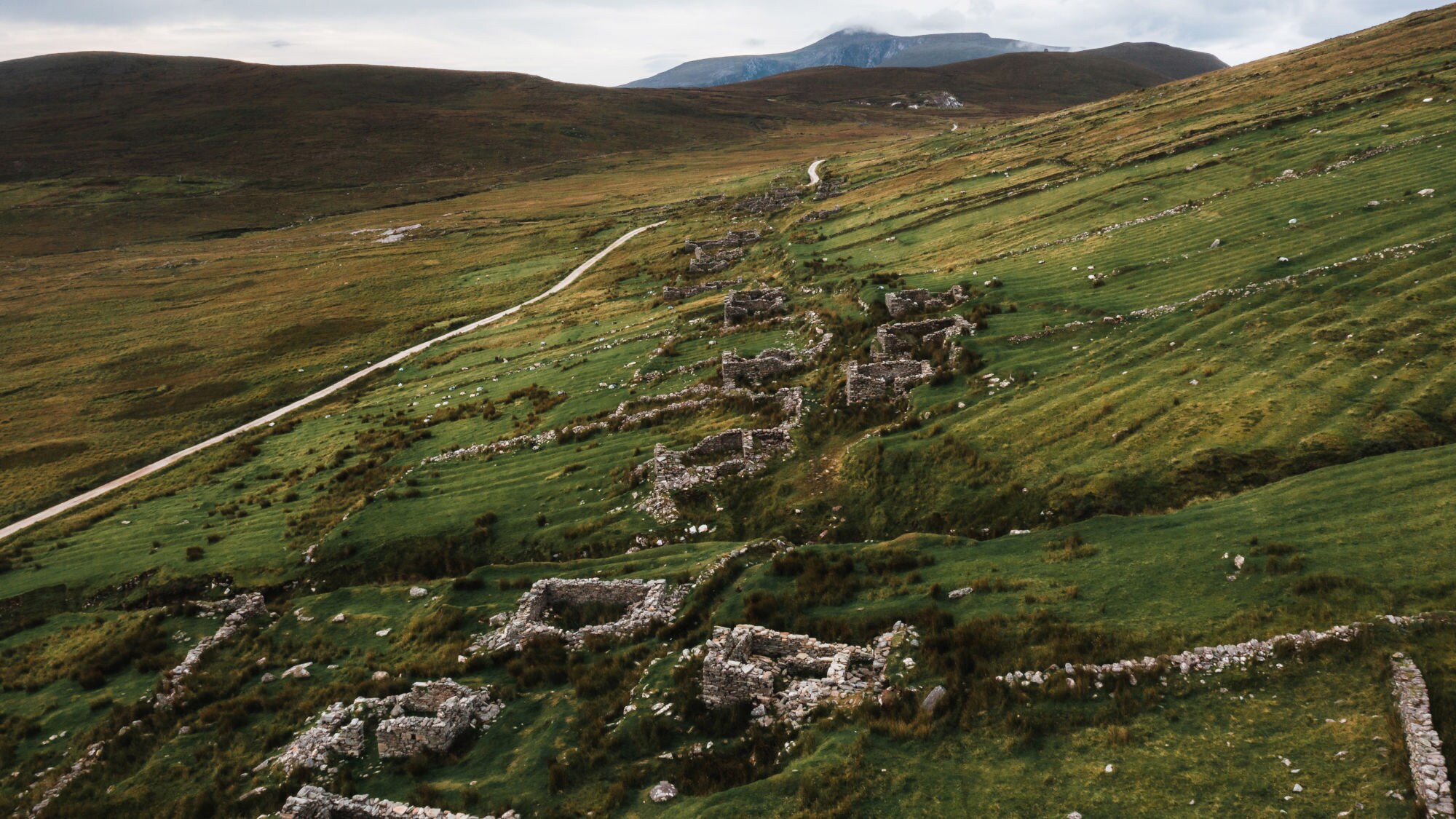 Landschaft mit grünen Wiesen und Überresten von Steinhäusern, die sich entlang eines Pfades erstrecken. Im Hintergrund sind sanfte Hügel sichtbar. Landschaft mit grünen Wiesen und Überresten von Steinhäusern, die sich entlang eines Pfades erstrecken. Im Hintergrund sind sanfte Hügel sichtbar.