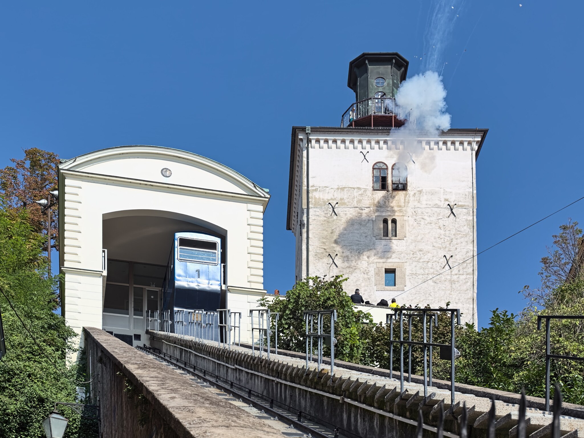 Weißer Turm mit Dampf auf dem Dach aus einer Kanone neben einer Standseilbahnstation auf einem Hügel.