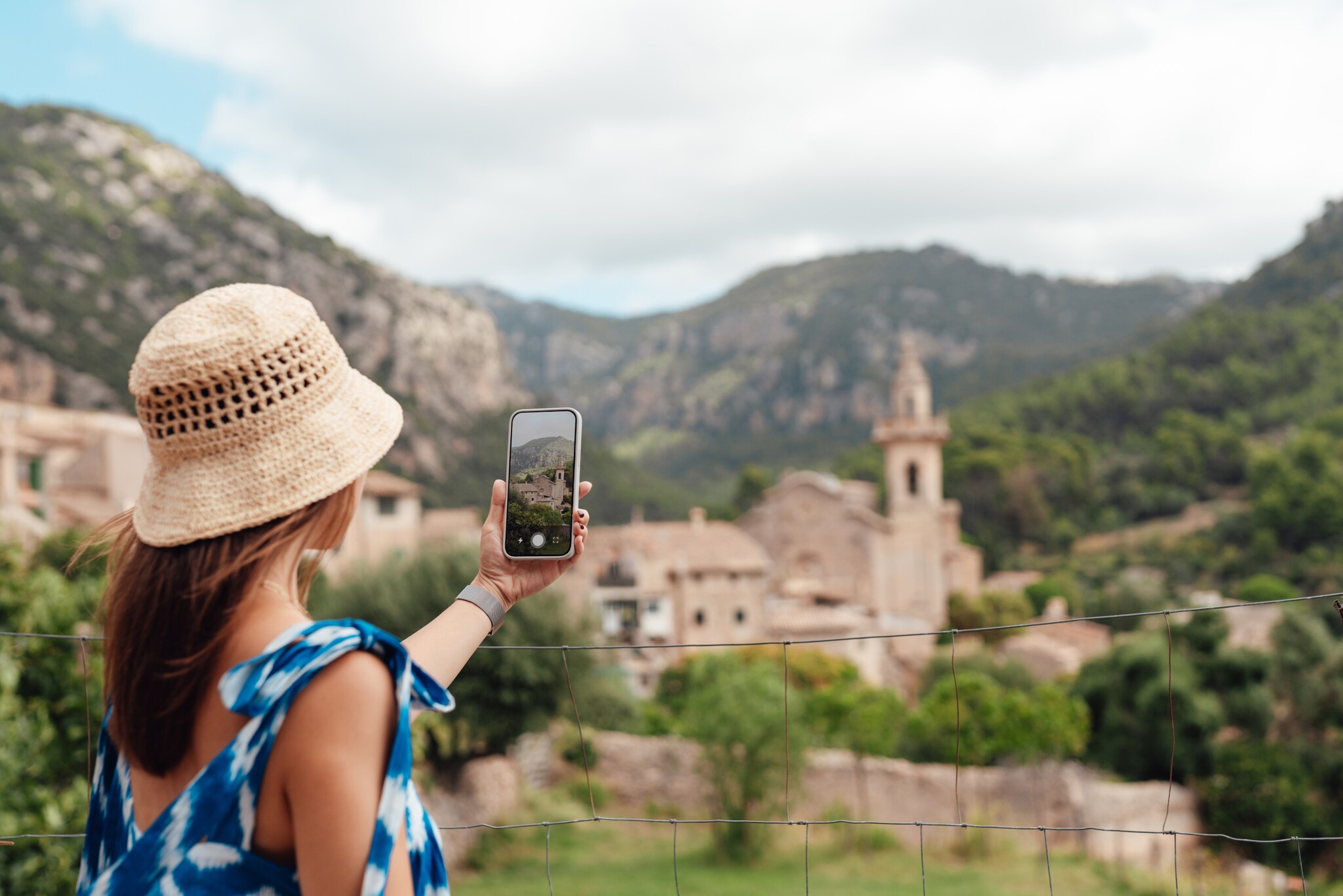 Eine Frau mit Strohhut und blauem Kleid fotografiert ein Bergdorf mit einem Smartphone. Eine Frau mit Strohhut und blauem Kleid fotografiert ein Bergdorf mit einem Smartphone.