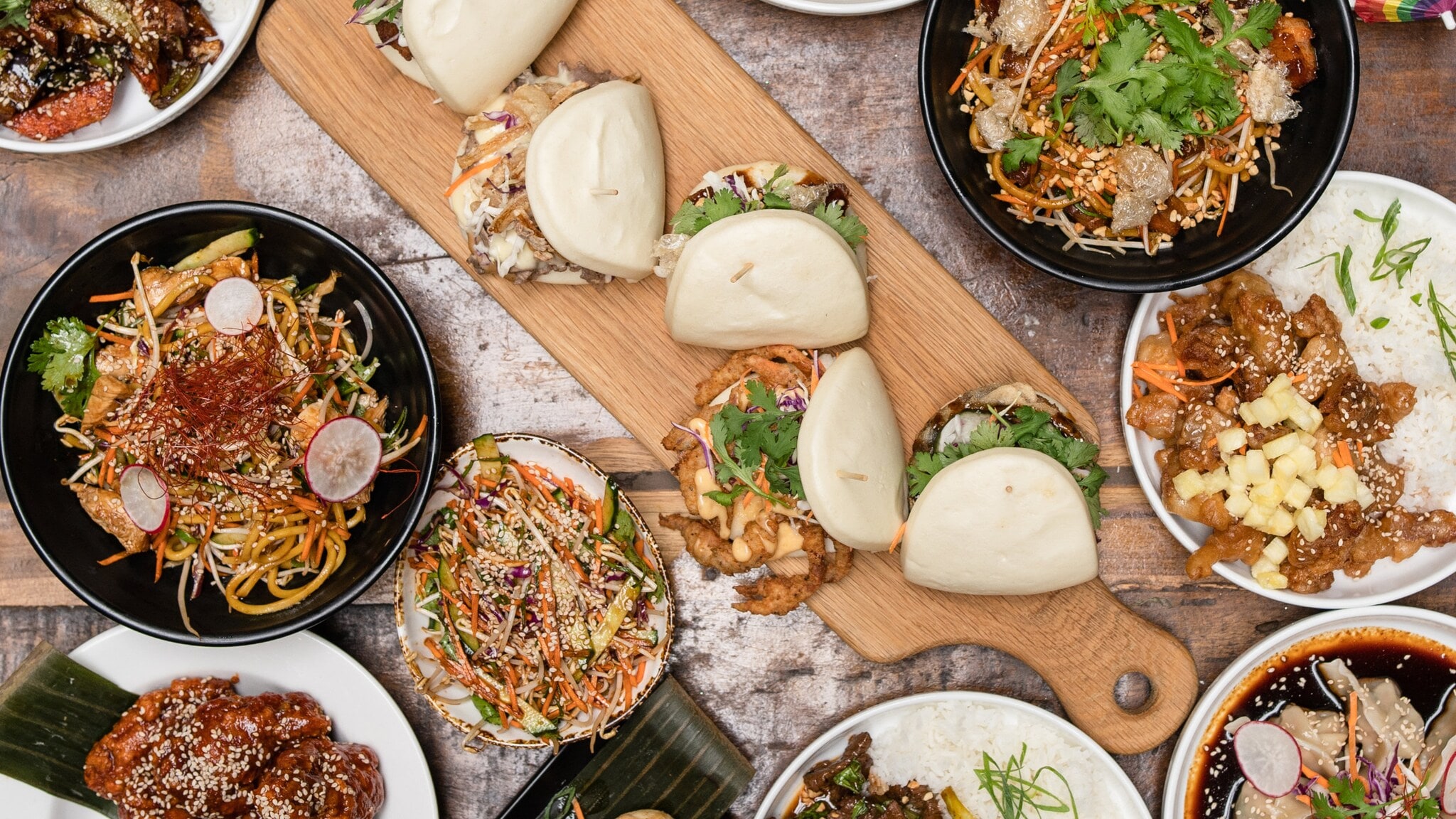 Eine Vielzahl von Gerichten auf einem Tisch: zwei Schalen mit gebratenen Nudeln, drei Schalen mit verschiedenen Fleischgerichten, Bao-Brötchen, Frühlingsrollen und Desserts. Eine Vielzahl von Gerichten auf einem Tisch: zwei Schalen mit gebratenen Nudeln, drei Schalen mit verschiedenen Fleischgerichten, Bao-Brötchen, Frühlingsrollen und Desserts.