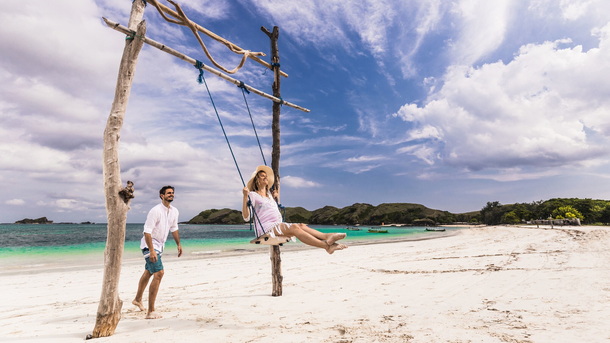 Ein Mann steht hinter einer schaukelnden schwangeren Frau auf einer Holzschaukel an einem weißen Sandstrand am türkisblauen Meer.
