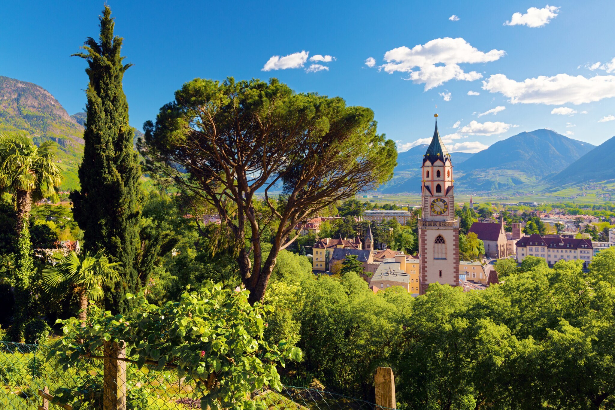 Panorama der Stadt Meran mit Kirchturm und üppiger Begrünung vor blauem Himmel und Gebirgszug am Horizont Panorama der Stadt Meran mit Kirchturm und üppiger Begrünung vor blauem Himmel und Gebirgszug am Horizont