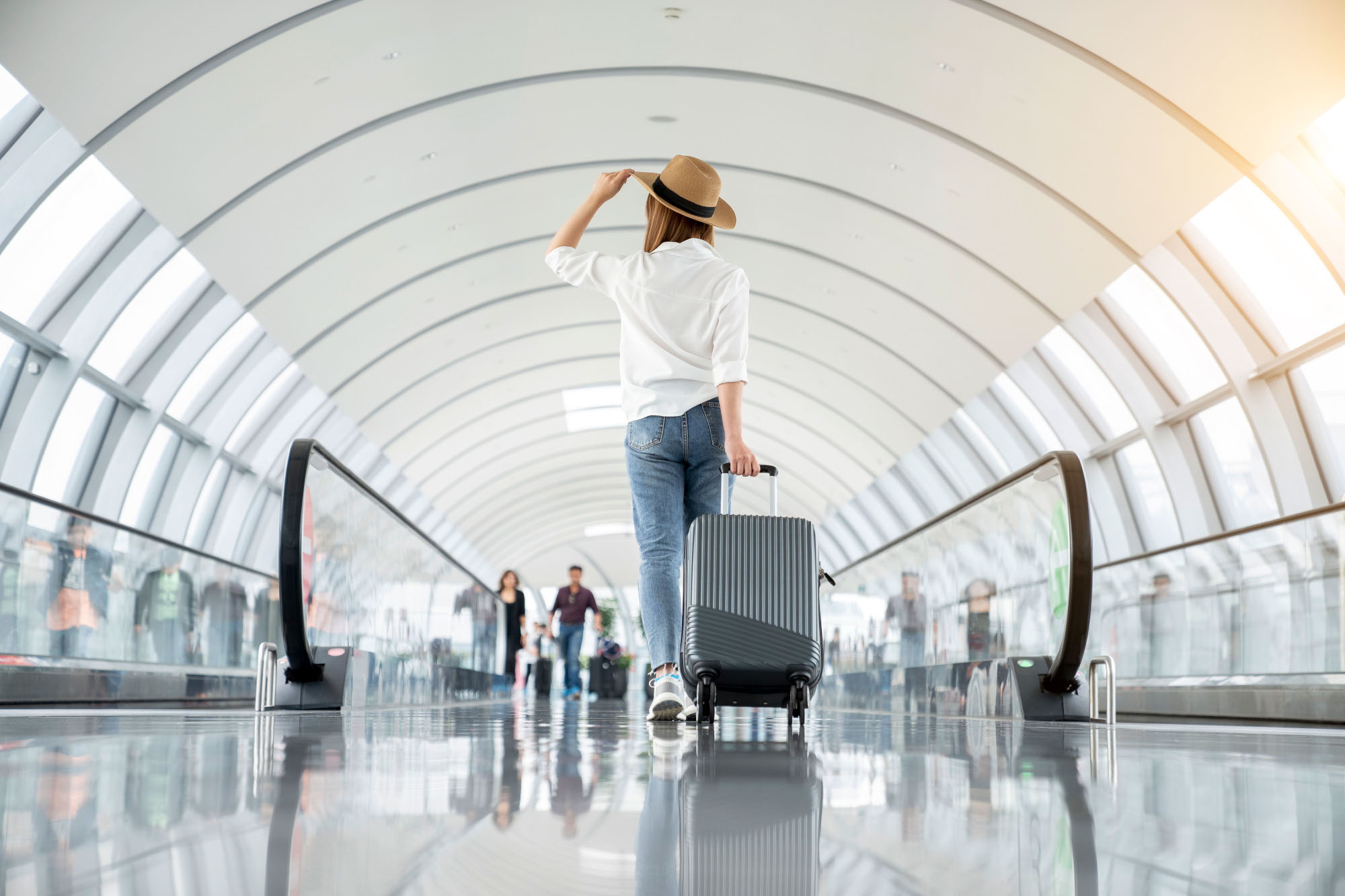 Rückansicht einer jungen Frau mit Strohhut, die in einem Flughafenterminal einen Trolley hinters ich herzieht.