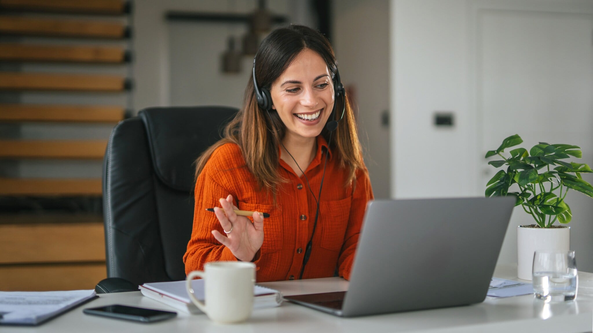 Eine Frau mit Headset winkt freundlich in Richtung Laptop vor ihr auf einem Tisch. Eine Frau mit Headset winkt freundlich in Richtung Laptop vor ihr auf einem Tisch.