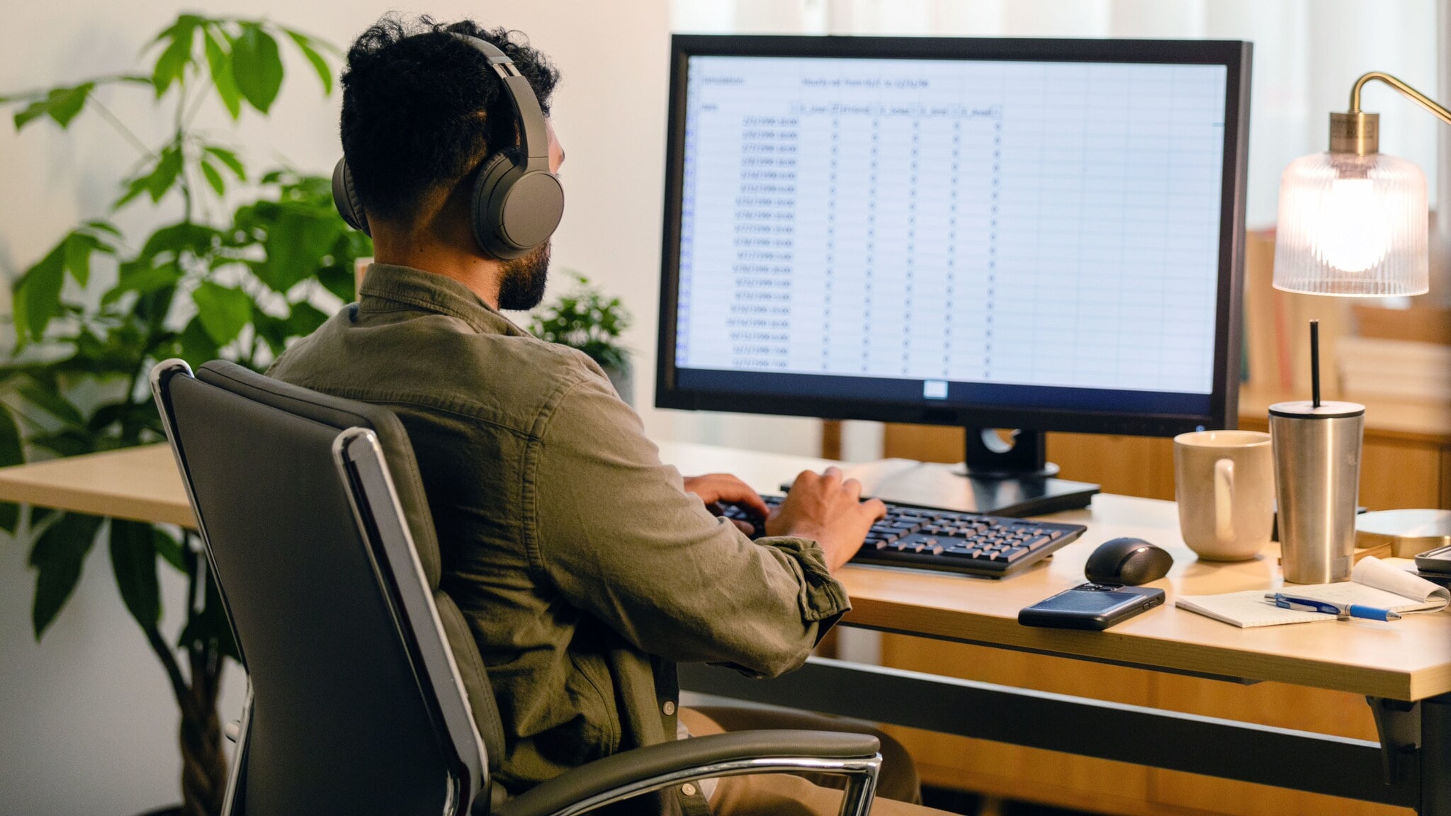 Rückansicht eines Mannes auf einem Bürostuhl, der mit Kopfhörern an einem Computer mit großem Display an einem Schreibtisch sitzt.