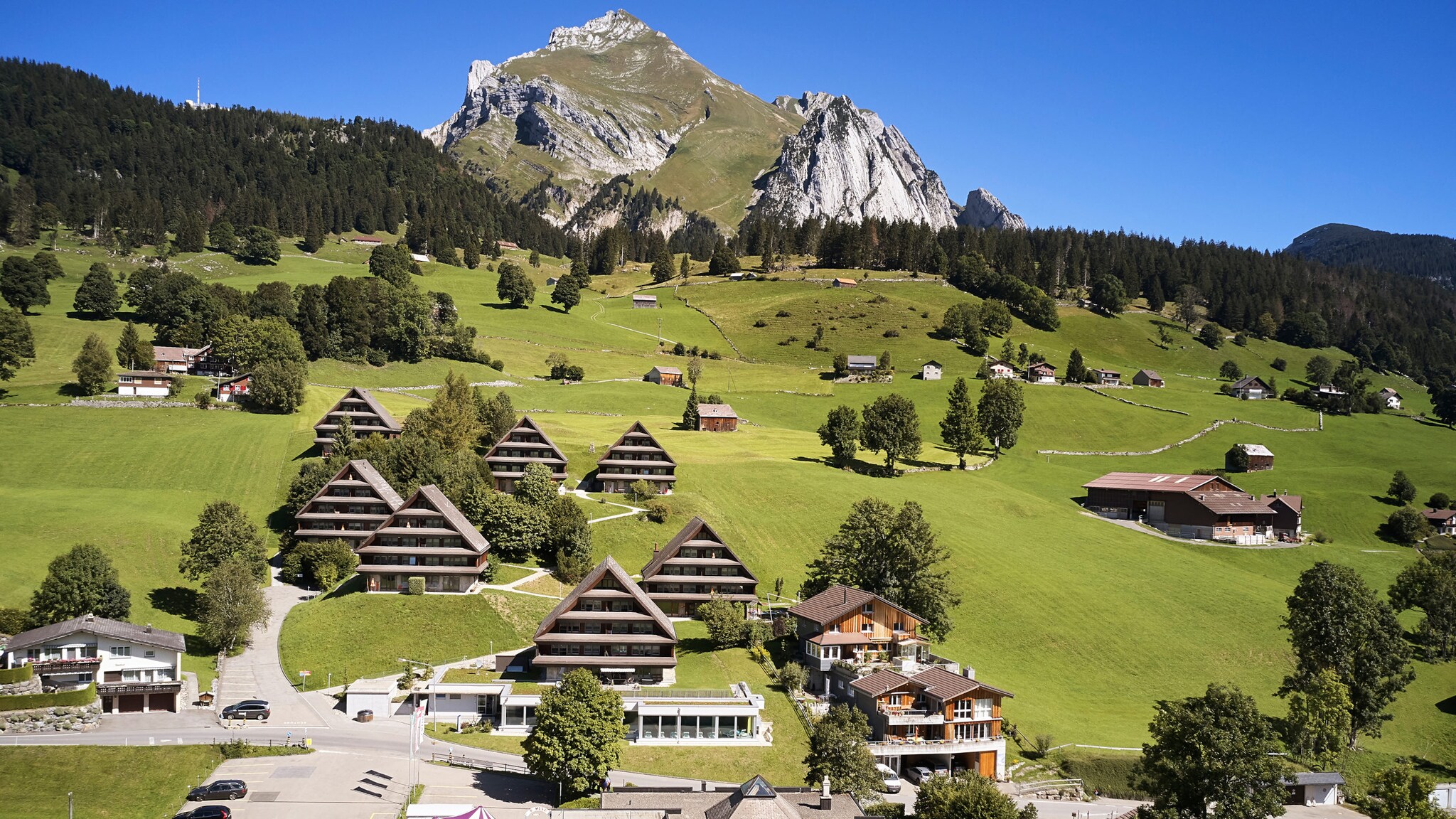Berglandschaft mit schneebedeckten Gipfeln, grünen Wiesen und verstreuten Bauernhäusern im Vordergrund. Wolken am Himmel.