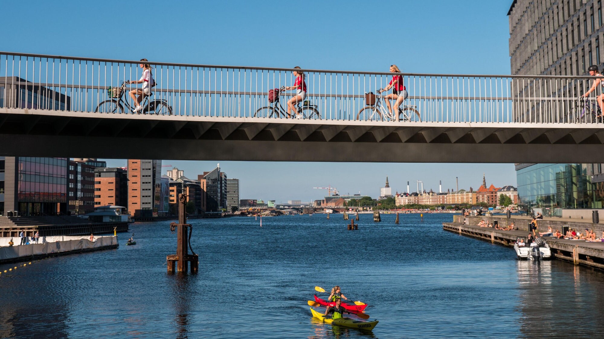 Mehrere Personen auf Fahrrädern fahren über eine Brücke über einem Fluss im Stadtzentrum im Sommer.