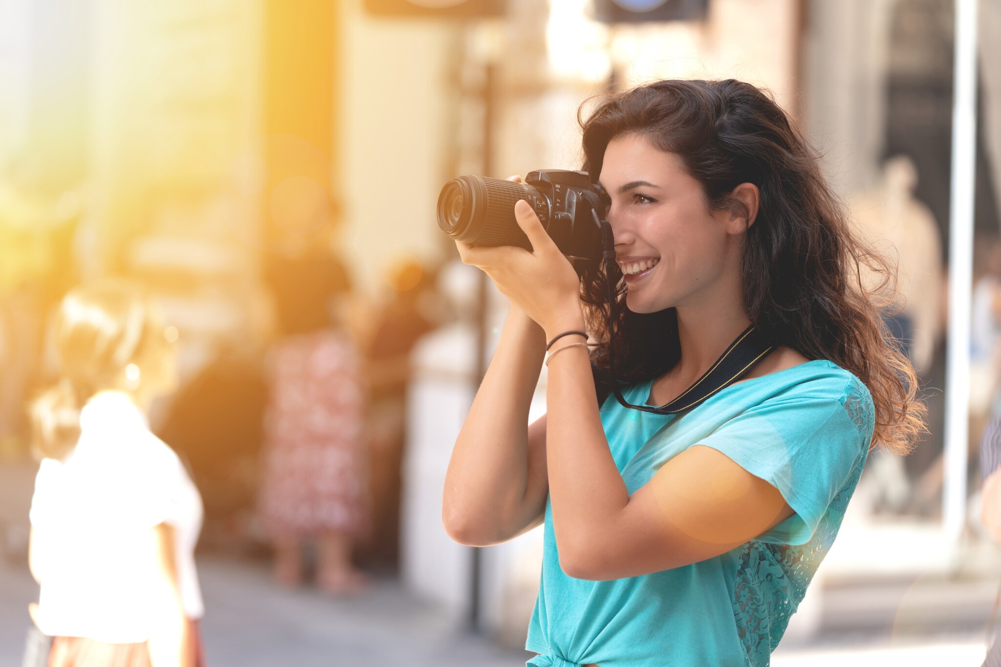 In einer urbanen Umgebung hält eine Frau in einem mintfarbenen T-Shirt eine Kamera vors Gesicht und blickt durch die Linse.