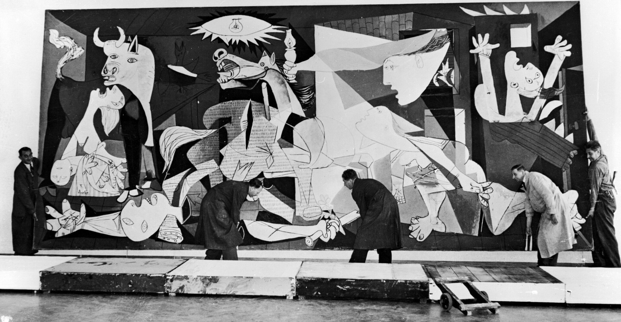 Historische Schwarzweißaufnahme von Picassos Monumentalgemäldes Guernica, das von vier Männern von der Wand genommen wird Historische Schwarzweißaufnahme von Picassos Monumentalgemäldes Guernica, das von vier Männern von der Wand genommen wird