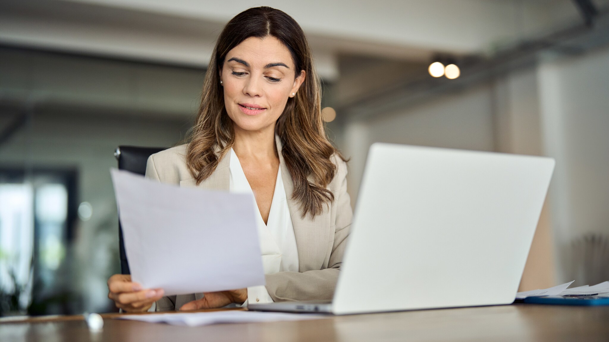 Eine Frau in Business-Kleidung sitzt an einem Schreibtisch an einem Laptop und schaut auf ein Papierblatt in ihrer Hand.