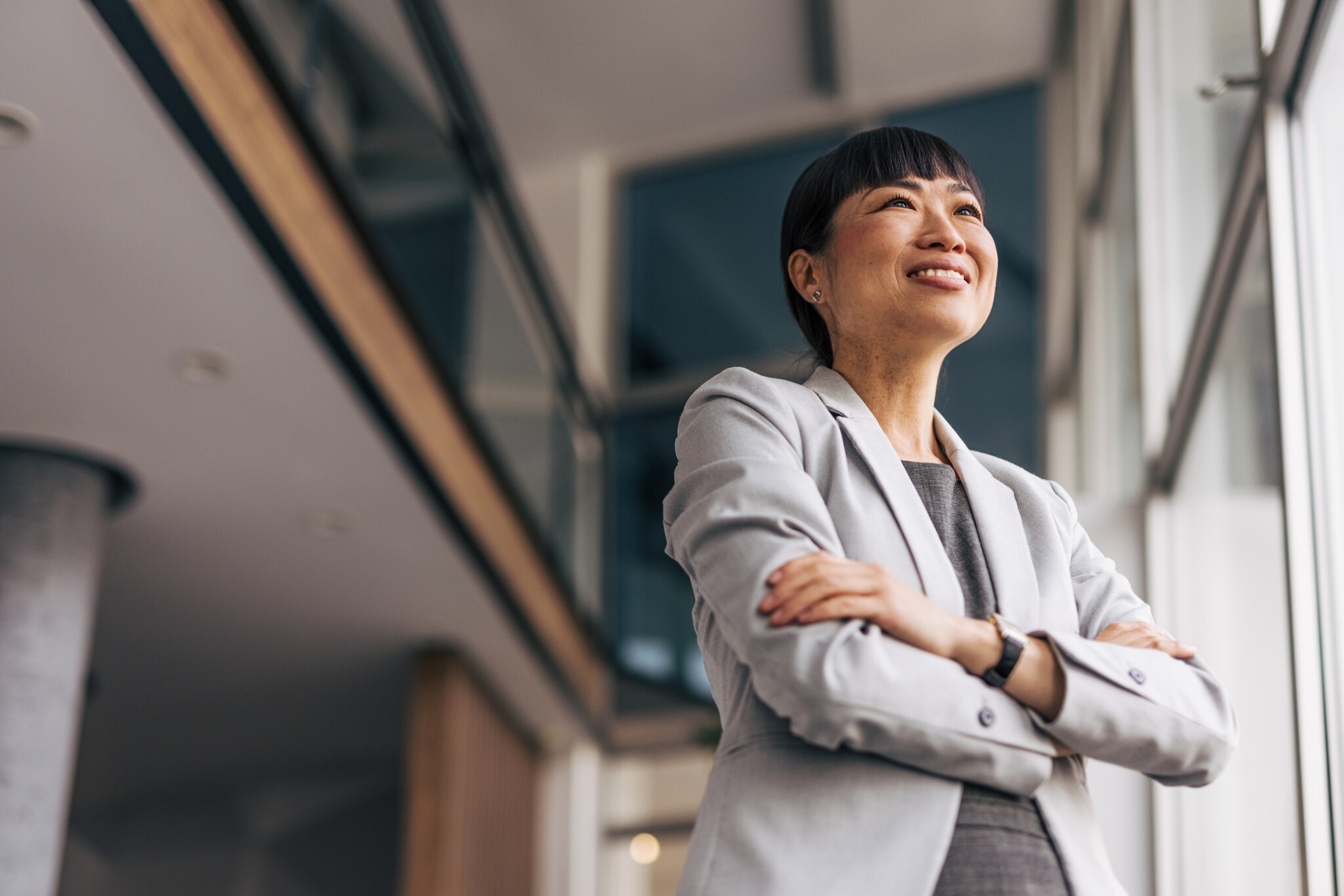 Porträt einer lächelnden Frau im Blazer mit verschränkten Armen in einem Bürogebäude mit viel Glas.