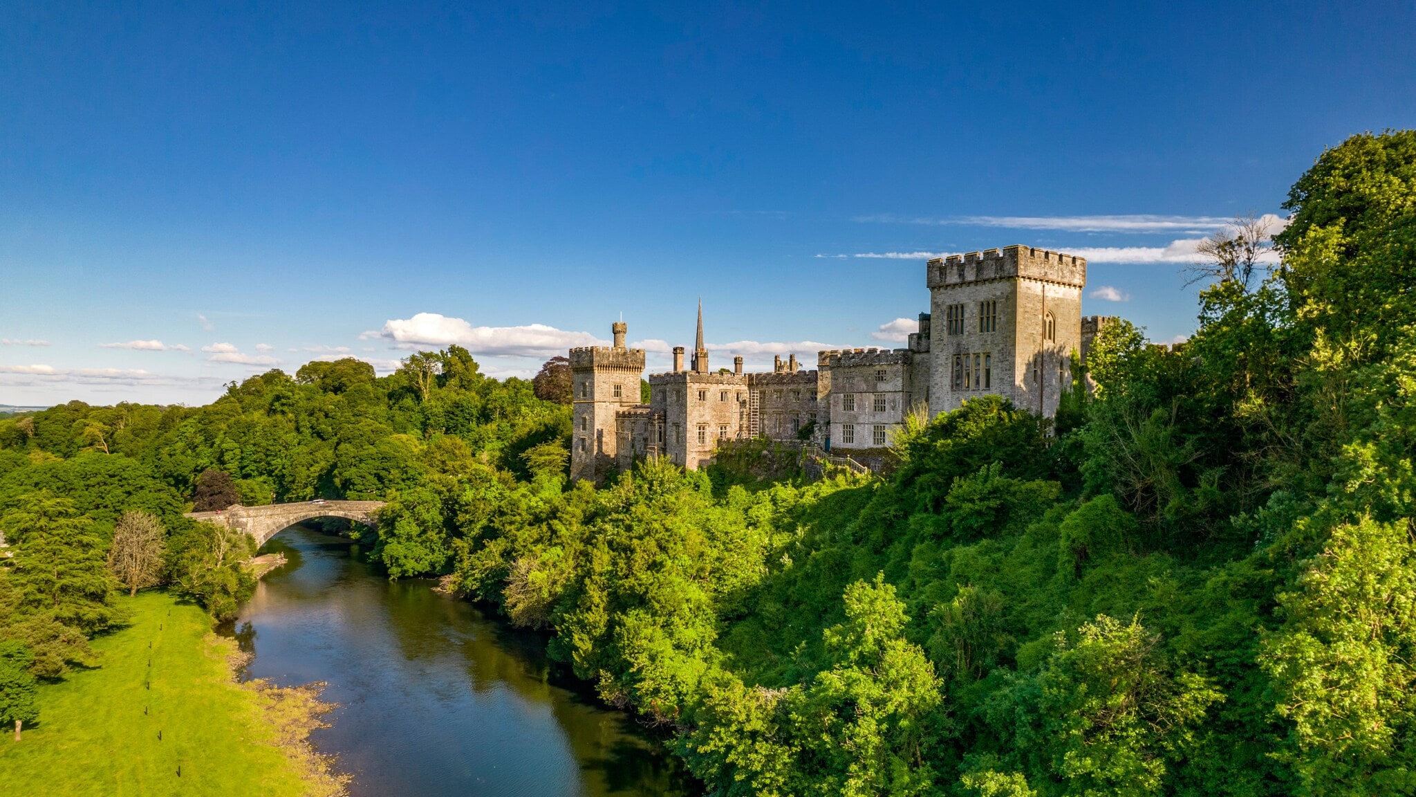 Luftaufnahme von Lismore Castle in Waterford, umgeben von dichtem Grün und einem Fluss mit einer Steinbrücke im Vordergrund. Luftaufnahme von Lismore Castle in Waterford, umgeben von dichtem Grün und einem Fluss mit einer Steinbrücke im Vordergrund.
