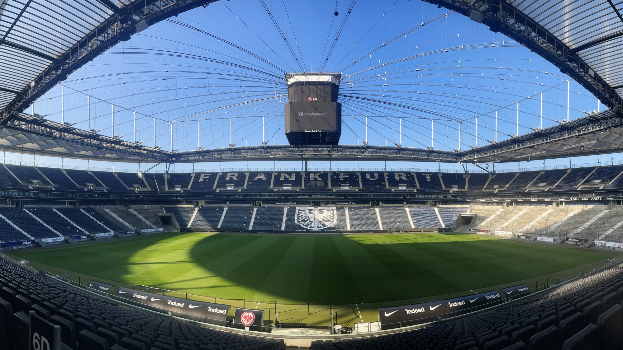 Innenansicht eines Fußballstadions mit Schriftzug Frankfurt und Adler-Wappen auf menschenleeren Rängen unter blauem Himmel.
