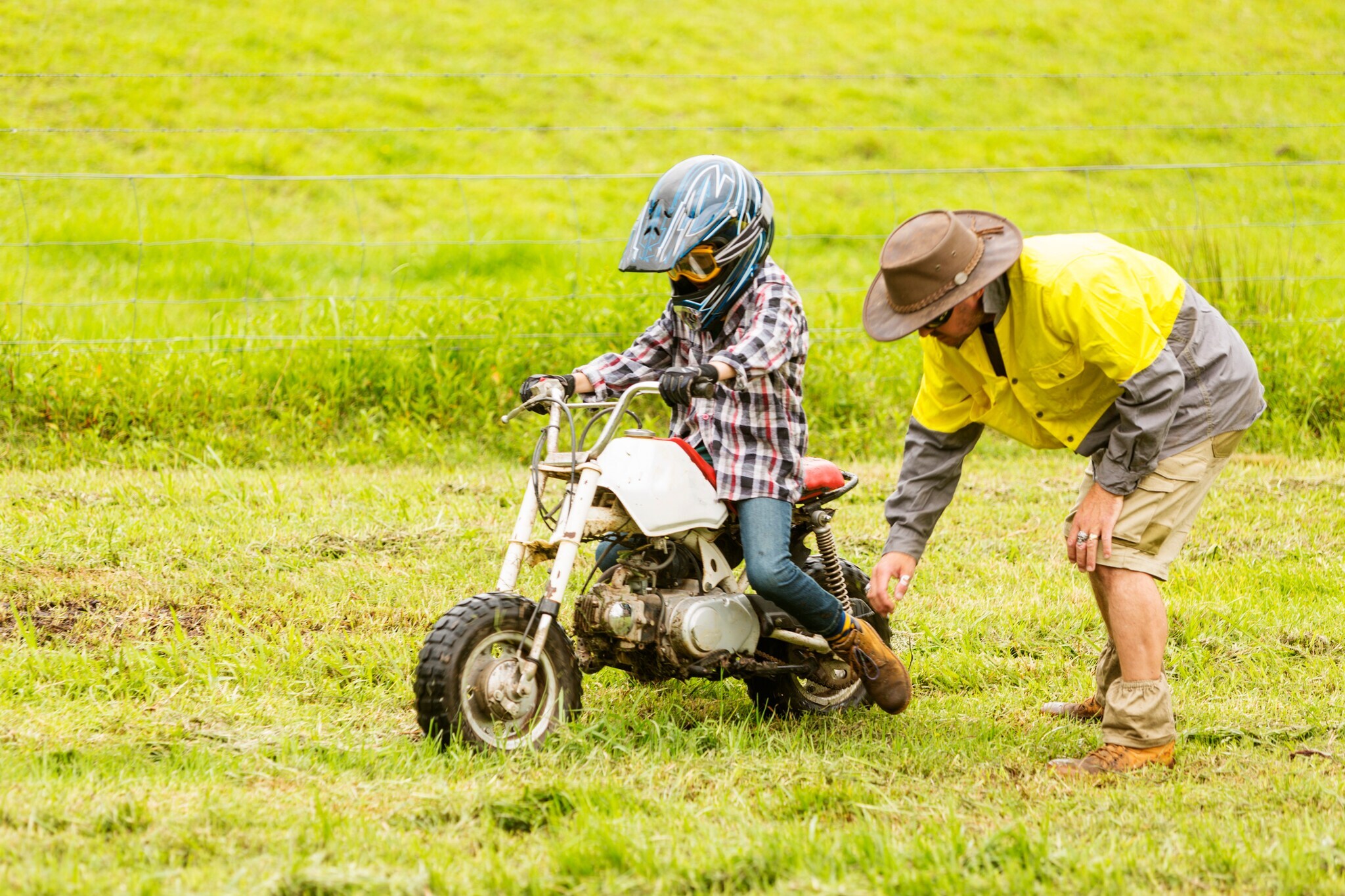 Kind auf Kindermotorrad auf einer feuchten Wiese, daneben ein gebückter Mann.