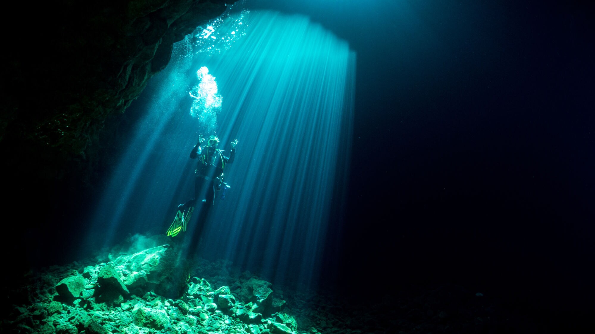 Ein Taucher schwebt in einem dunklen Unterwasserraum, umgeben von Lichtstrahlen, die von der Oberfläche kommen. Der Boden besteht aus Steinen.