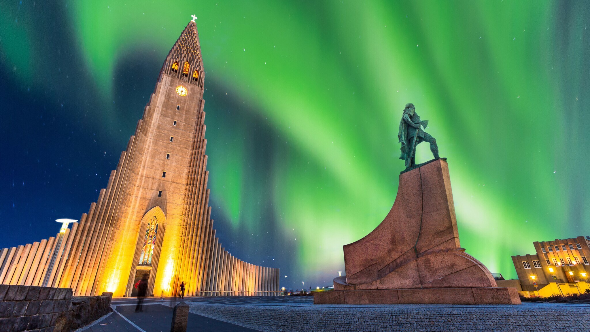 Kirche und Statue aus der Untersicht auf einem Platz vor Nachthimmel mit grün leuchtenden Polarlichtern.