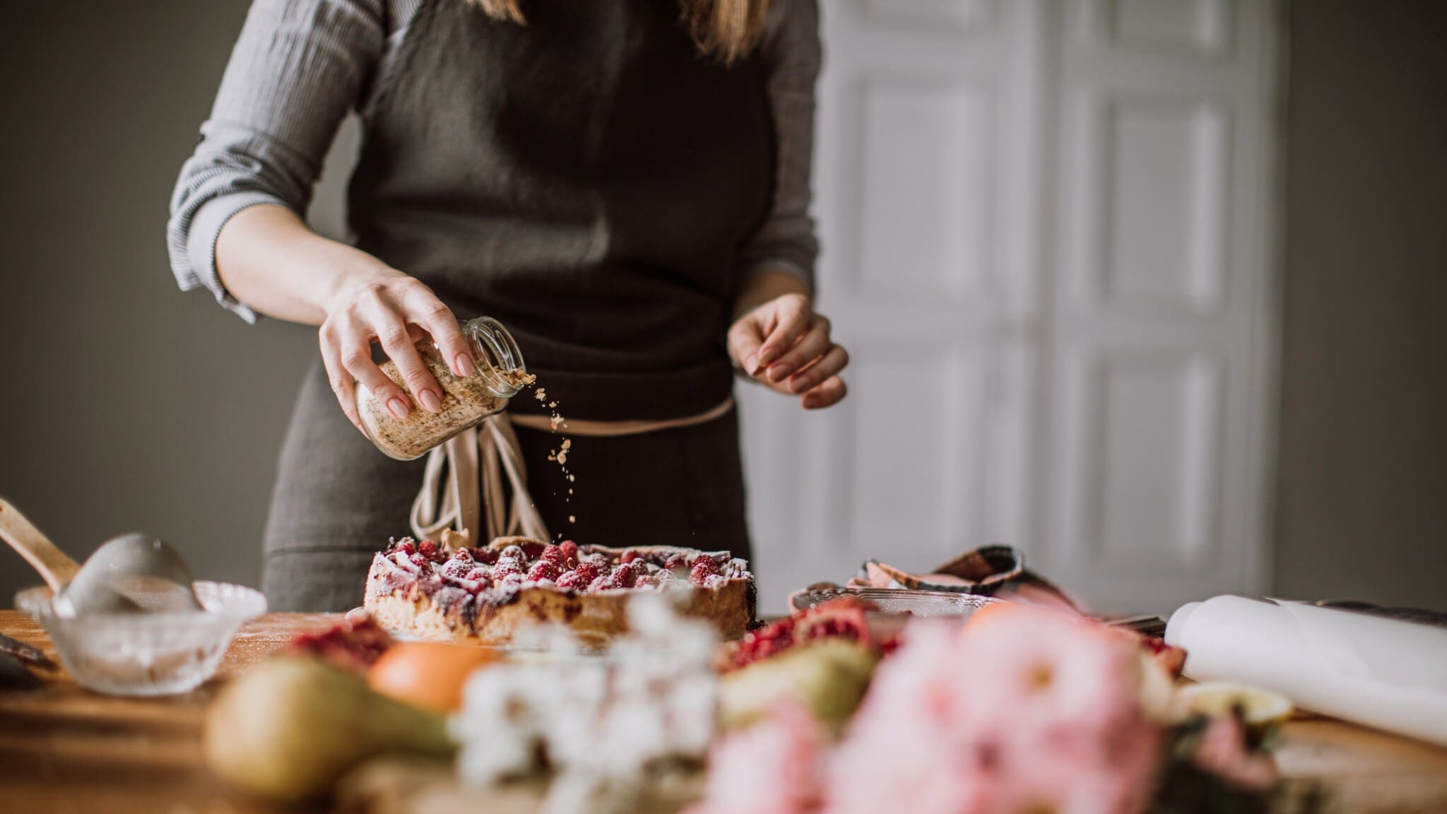 Eine Hand streut Streusel aus einem Glas auf einen Kuchen, der mit Himbeeren dekoriert ist und auf einem Tisch steht.