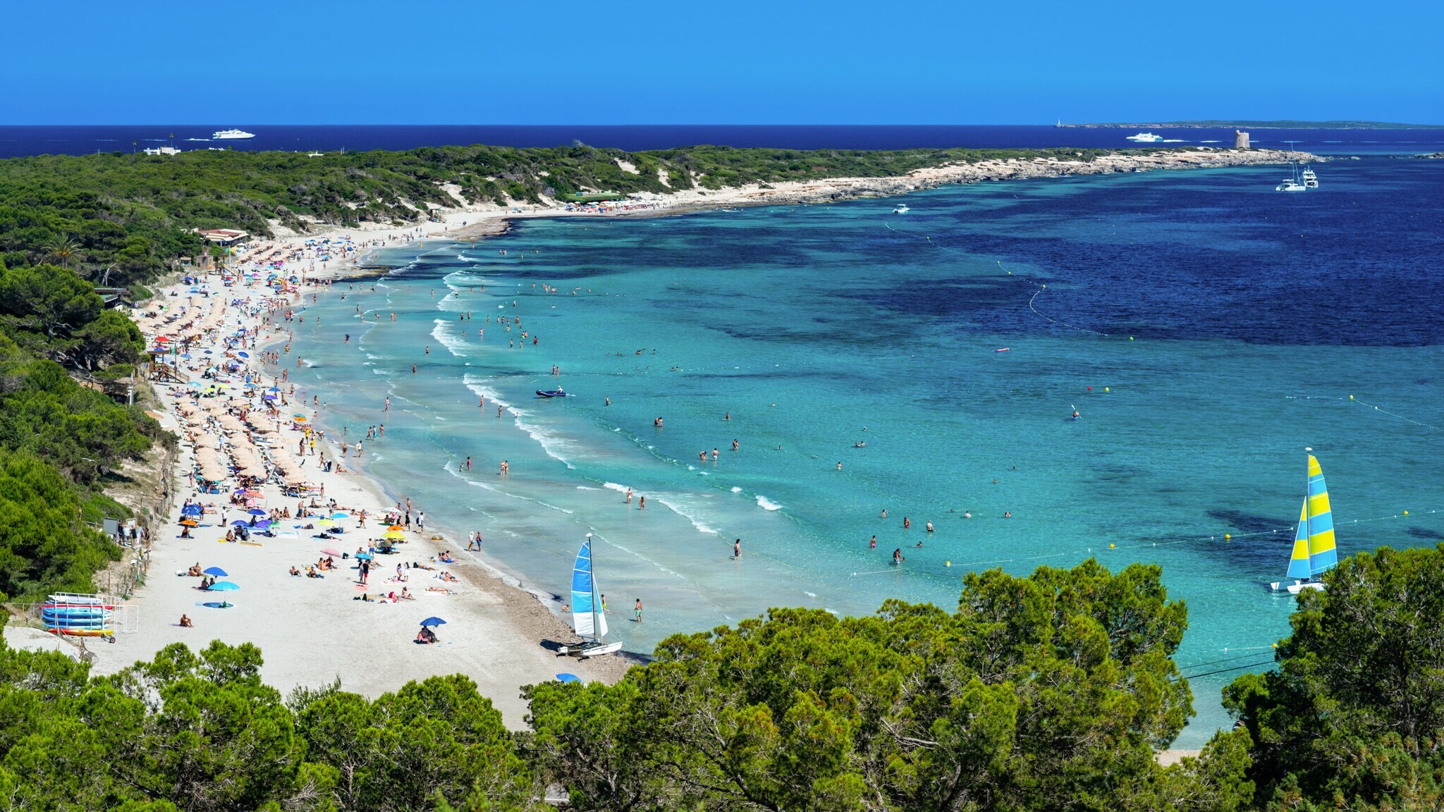 Strand mit vielen Menschen, türkisfarbenem Wasser und Segelbooten an der Küste, umgeben von grünen Bäumen