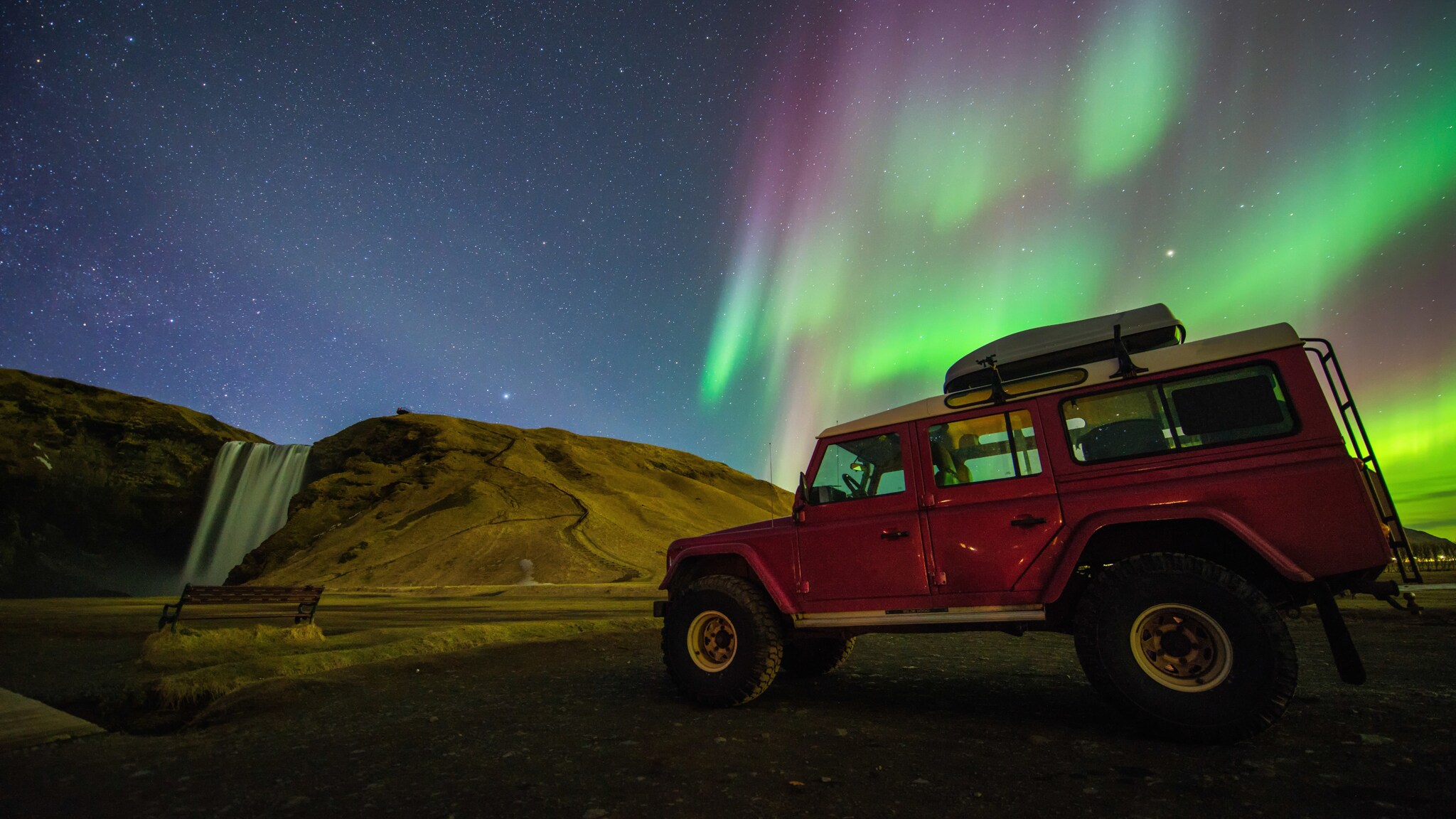 Ein roter Jeep parkt in einem Naturgebiet mit Wasserfall bei Polarlichtern am nächtlichen Sternenhimmel.