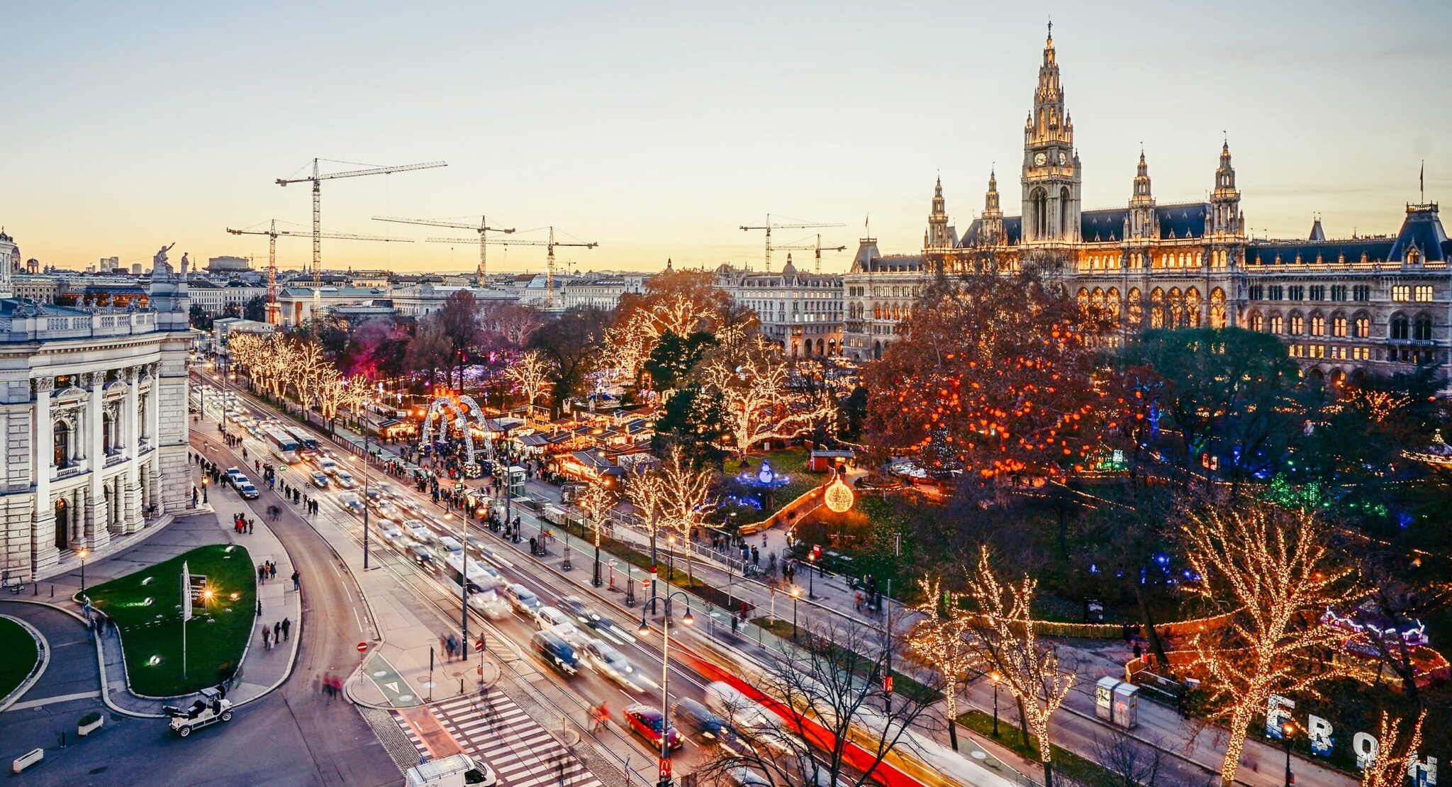 Panoramablick auf das beleuchtete Wiener Rathaus mit Weihnachtsmarkt auf dem großen Vorplatz, davor eine mehrspurige Straße