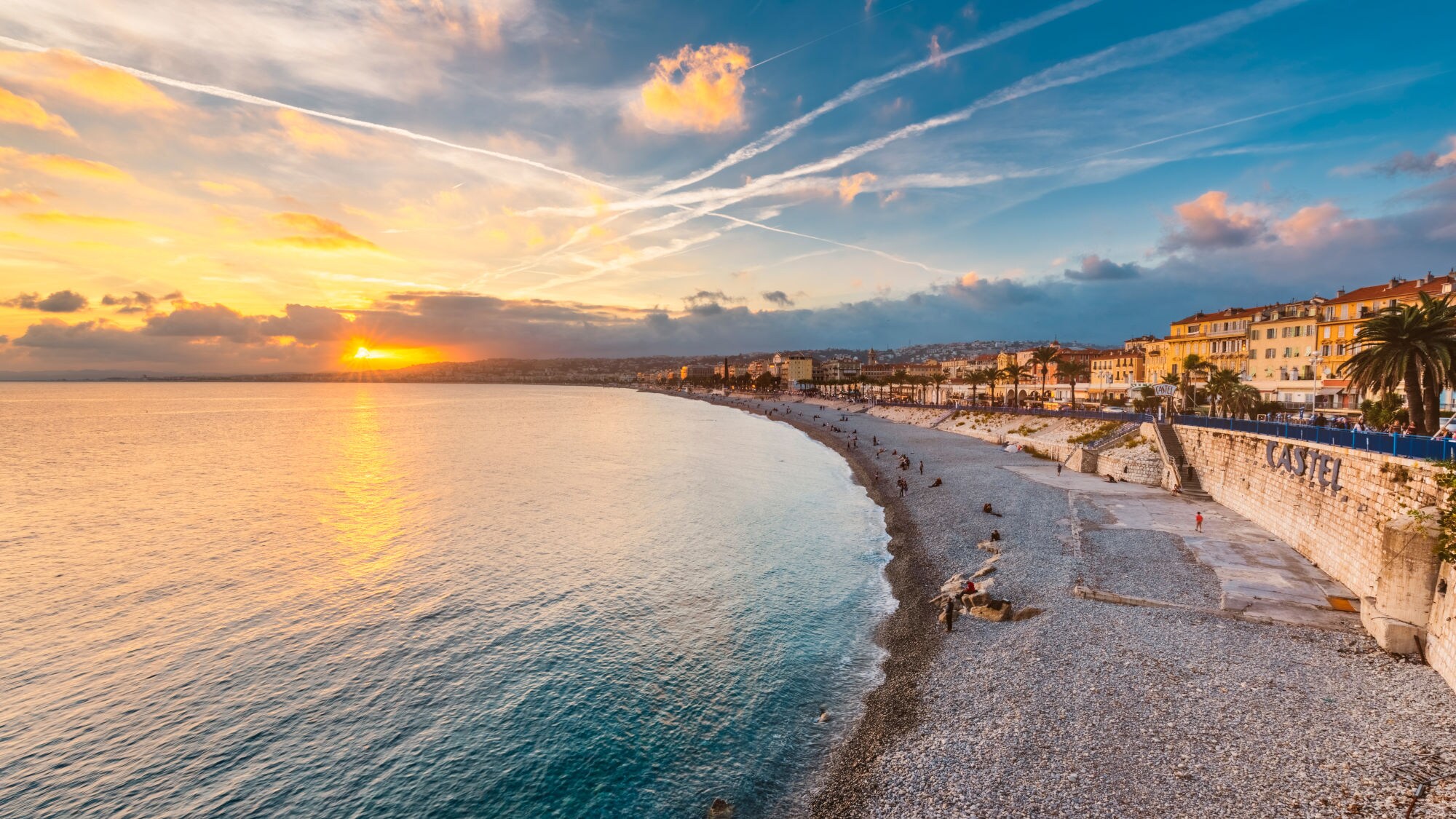 Blick auf die Küste von Nizza bei Sonnenuntergang, mit einem Kiesstrand, Palmen und Gebäuden entlang der Uferpromenade. Blick auf die Küste von Nizza bei Sonnenuntergang, mit einem Kiesstrand, Palmen und Gebäuden entlang der Uferpromenade.