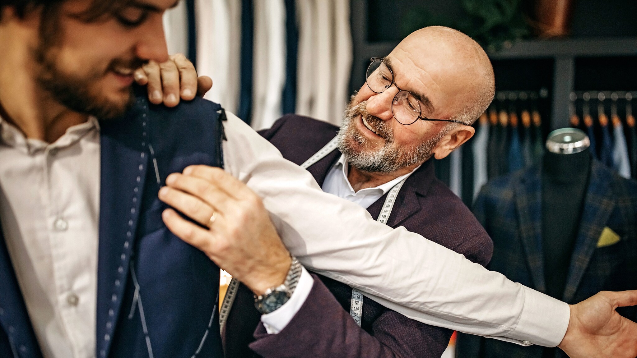 Ein Mann mit Maßband um den Hals fasst an die Schulter eines anderen Mannes, der den Arm ausstreckt.