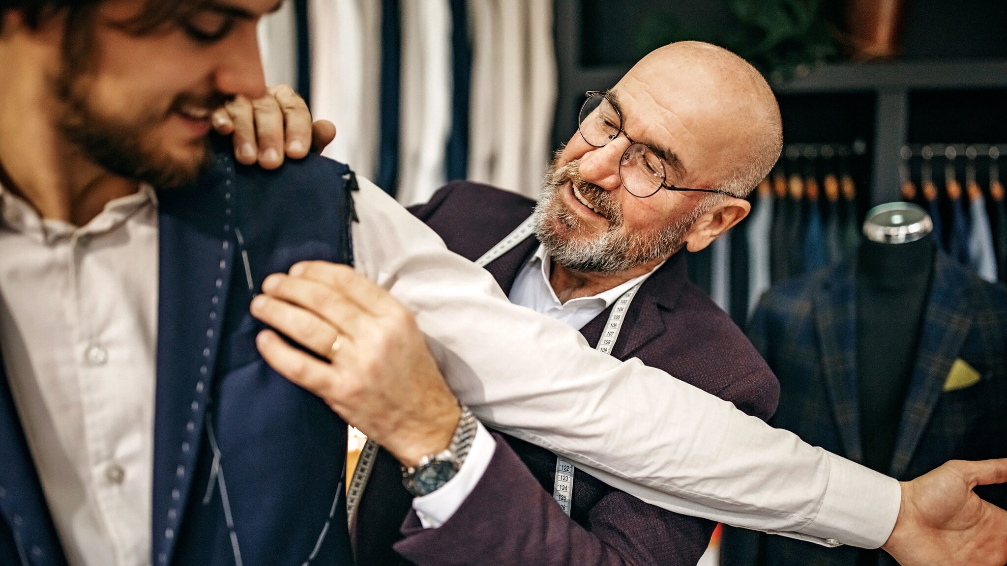 Ein Mann mit Maßband um den Hals fasst an die Schulter eines anderen Mannes, der den Arm ausstreckt. Ein Mann mit Maßband um den Hals fasst an die Schulter eines anderen Mannes, der den Arm ausstreckt.