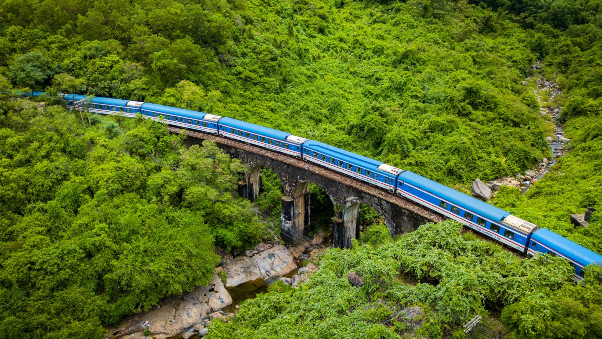 Ein blauer Zug fährt über eine Brücke aus Stein, umgeben von dichtem, grünem Wald und Felsen. Im Vordergrund ist ein Flussbett sichtbar. Ein blauer Zug fährt über eine Brücke aus Stein, umgeben von dichtem, grünem Wald und Felsen. Im Vordergrund ist ein Flussbett sichtbar.