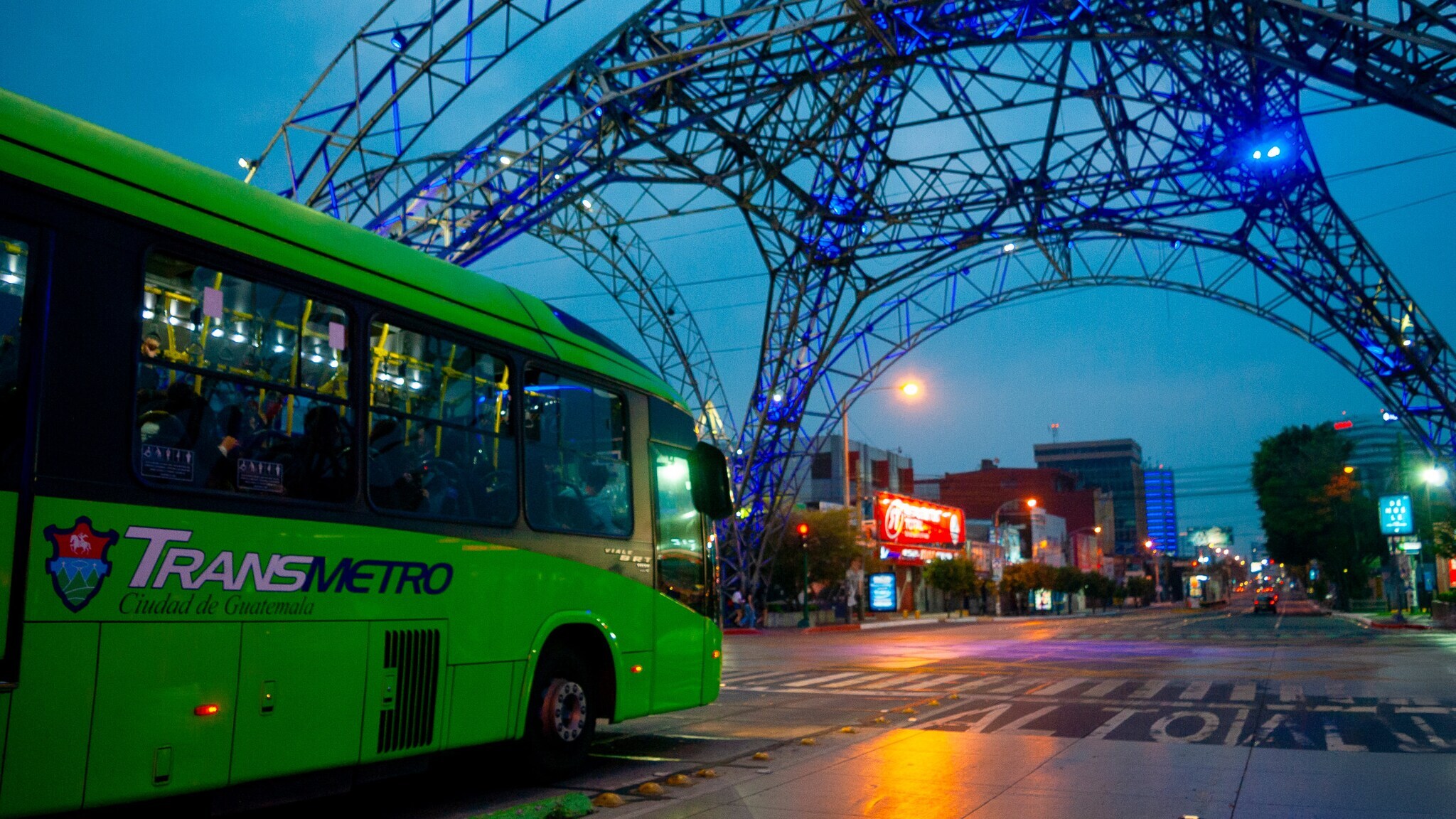 Grüner Transmetro-Bus unter beleuchtetem Metallbogen in der Stadt Guatemala bei Dämmerung