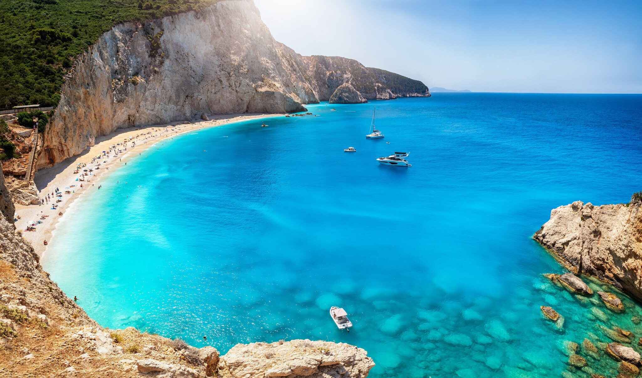 Blick von oben auf einen Sandstrand in einer von Felsen gesäumten Bucht am türkisblauen Meer.