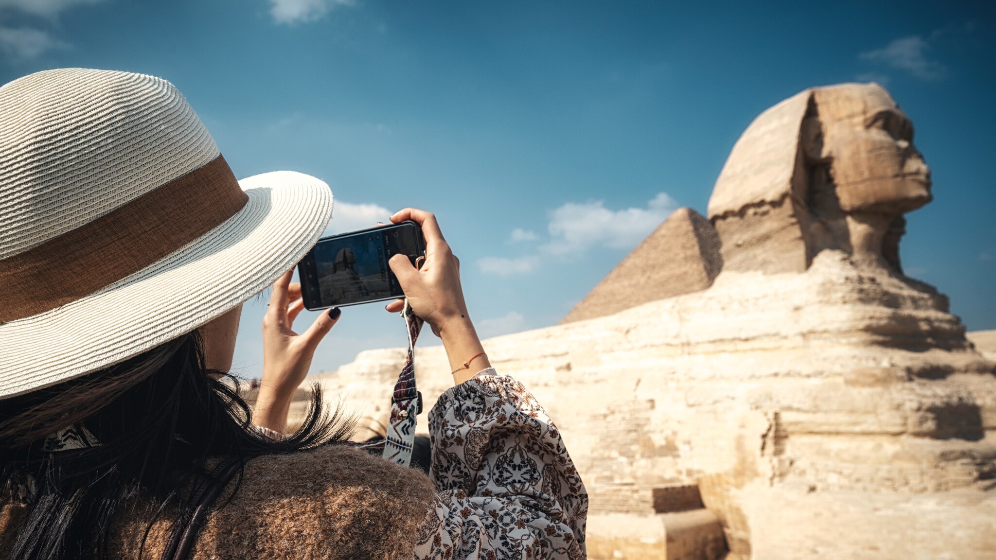 Eine Frau mit Hut fotografiert die Große Sphinx von Gizeh mit einem Smartphone bei klarem Himmel. Eine Frau mit Hut fotografiert die Große Sphinx von Gizeh mit einem Smartphone bei klarem Himmel.