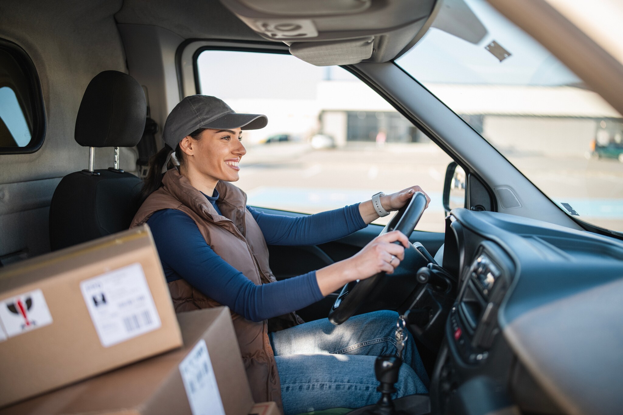 Eine lächelnde Frau mit Basecap sitzt am Steuer eines Transporters. Auf dem Beifahrersitz liegen gestapelte Kartons.
