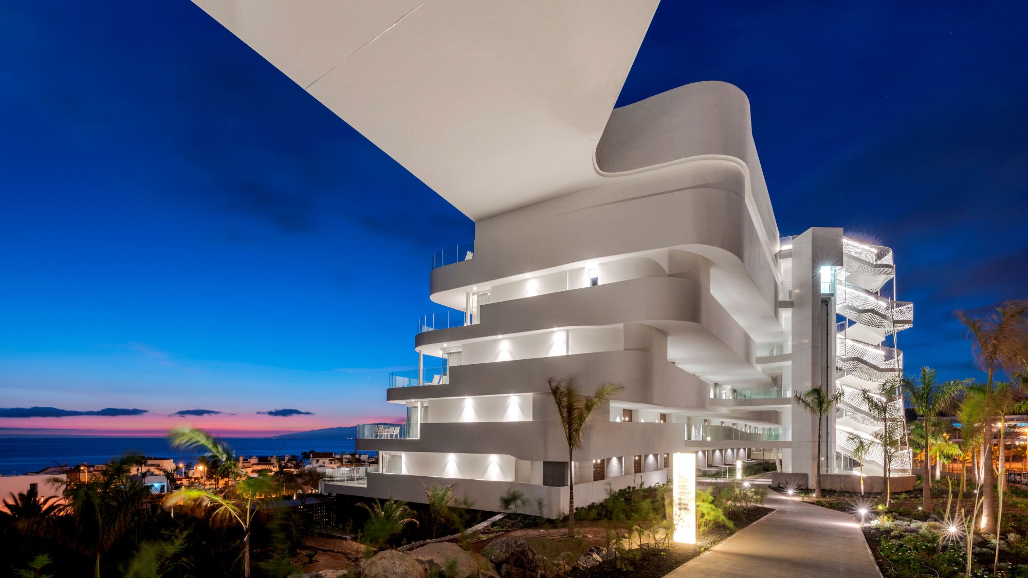 Modernes, weißes Gebäude mit geschwungenen Balkonen bei Nacht, beleuchtet, mit Blick auf das Meer und Palmen auf Teneriffa.