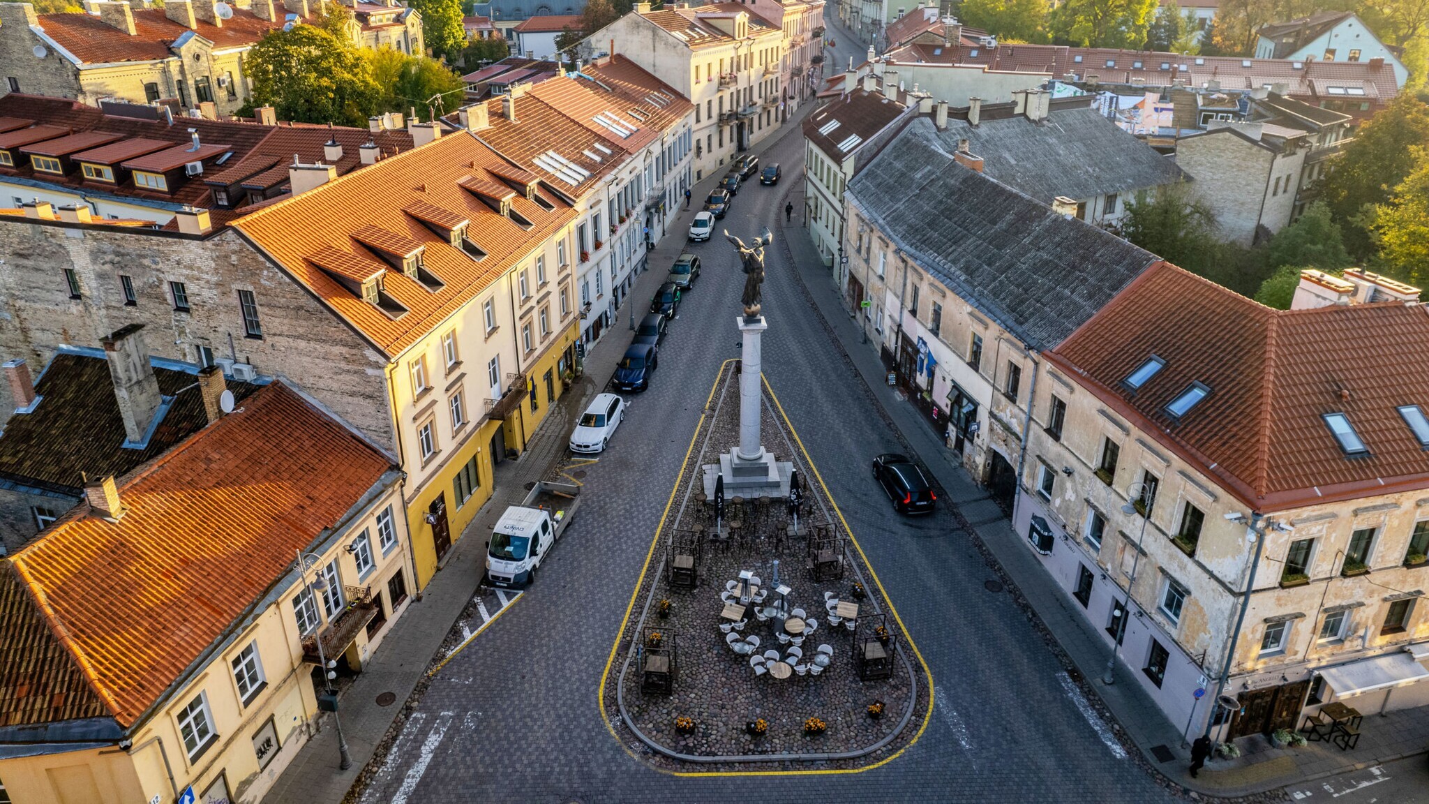Luftaufnahme eines Straßenzuges mit einer Verkehrsinsel mit Siegessäule und Sitzplätzen in einem Altstadtviertel.