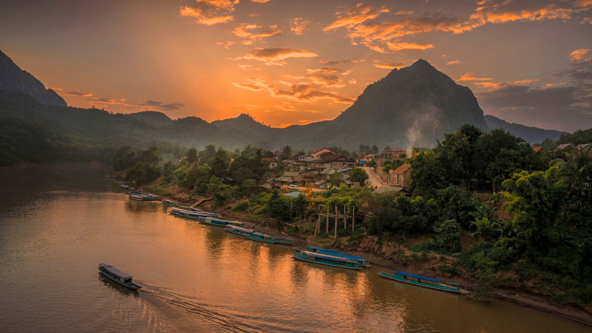 Landschaft in Laos mit einem Fluss, umgeben von Bergen und Dörfern. Der Himmel ist orange und rosa gefärbt, Boote liegen am Ufer.