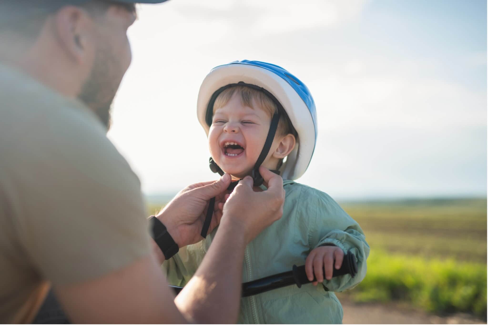 Kind mit blau-weißem Fahrradhelm hält Lenker eines Fahrrads, während eine erwachsene Person den Helm festzieht Kind mit blau-weißem Fahrradhelm hält Lenker eines Fahrrads, während eine erwachsene Person den Helm festzieht