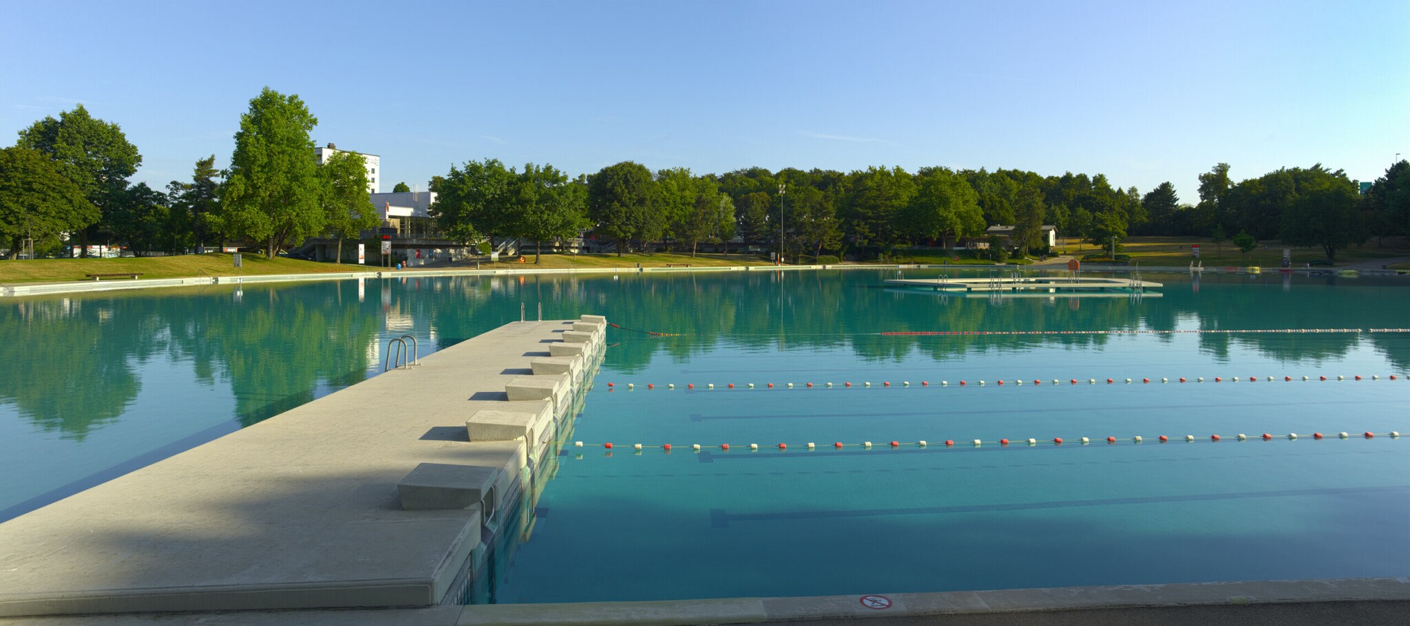 Ein Baggersee-ähnliches Schwimmbecken im Freien, mit abgetrennten Bahnen und einer schmalen, ringförmigen Kunstinsel.