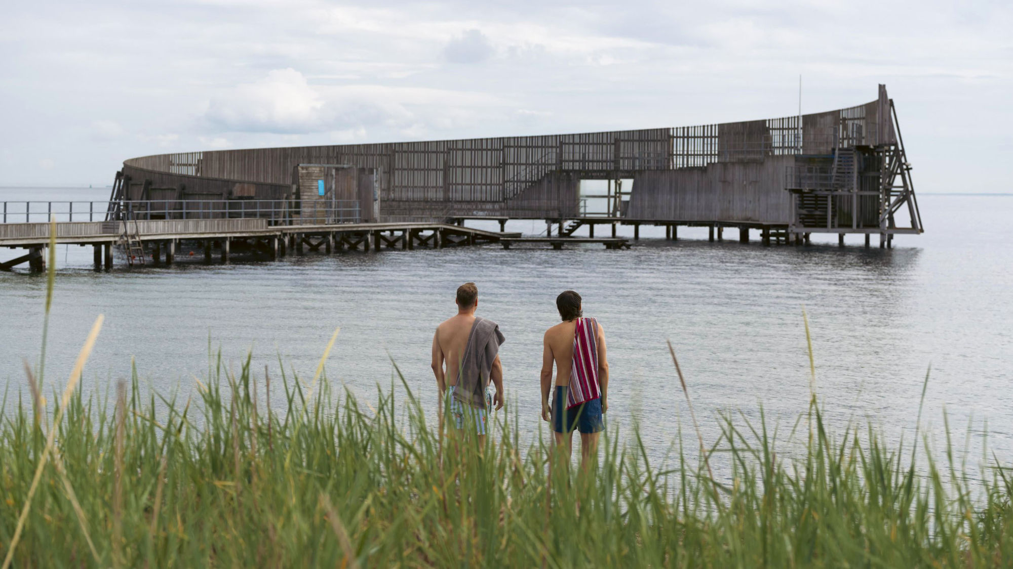 Rückansicht zweier Männer mit nackten Oberkörpern und Handtüchern über den Schultern vor einer Holzkonstruktion mit Steg im Meer.
