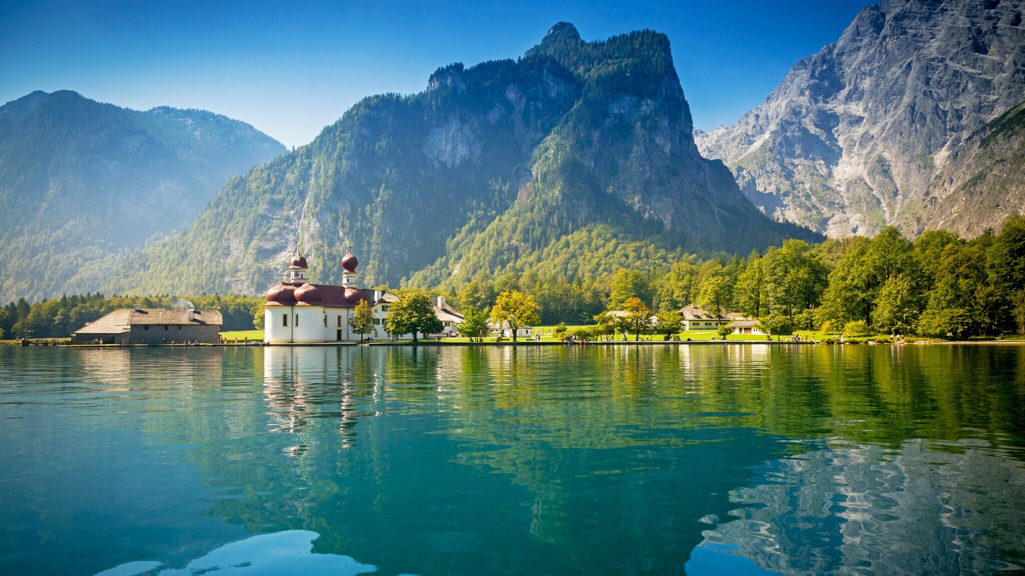 Kirche mit Zwiebeltürmen am Ufer eines klaren Sees, umgeben von bewaldeten Bergen unter blauem Himmel.