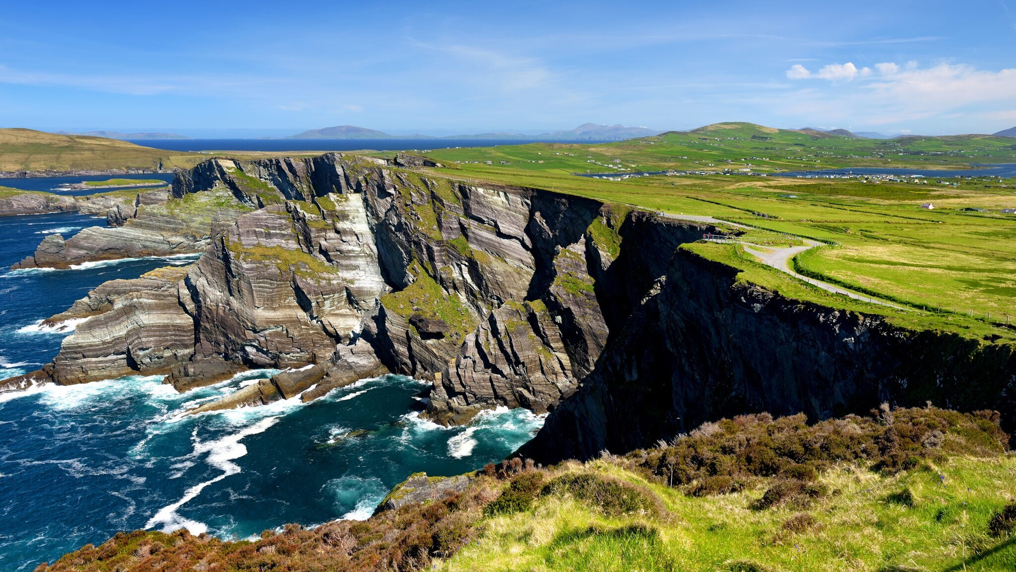 Luftaufnahme einer Küstenlandschaft mit steilen Klippen, grünem Gras und einem schmalen Weg, der entlang der Klippen verläuft. Im Hintergrund sind sanfte Hügel und ein blauer Himmel zu sehen.