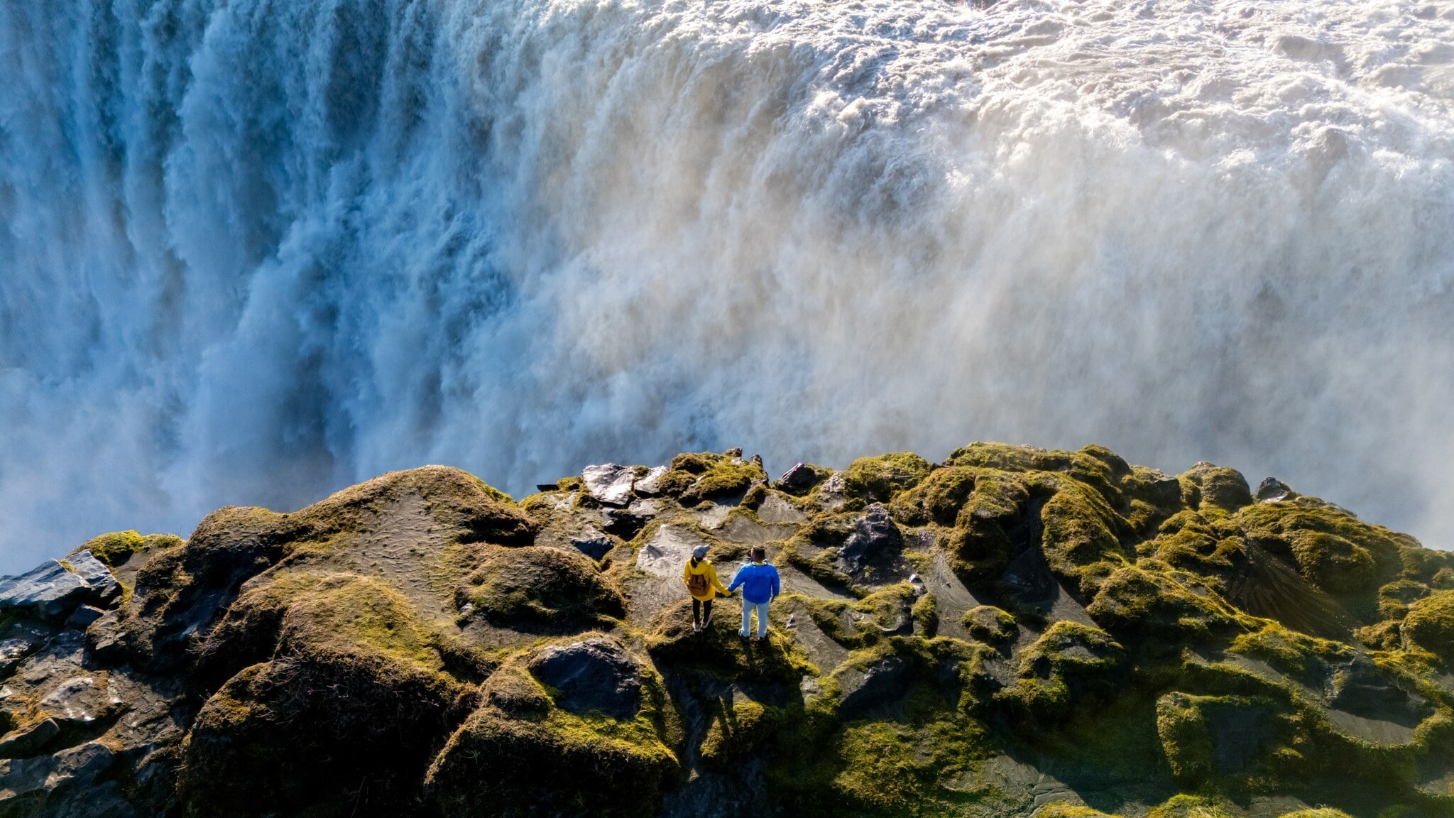 Zwei Personen stehen auf moosbedecktem Felsen vor einem großen Wasserfall mit starker Gischtentwicklung. Zwei Personen stehen auf moosbedecktem Felsen vor einem großen Wasserfall mit starker Gischtentwicklung.
