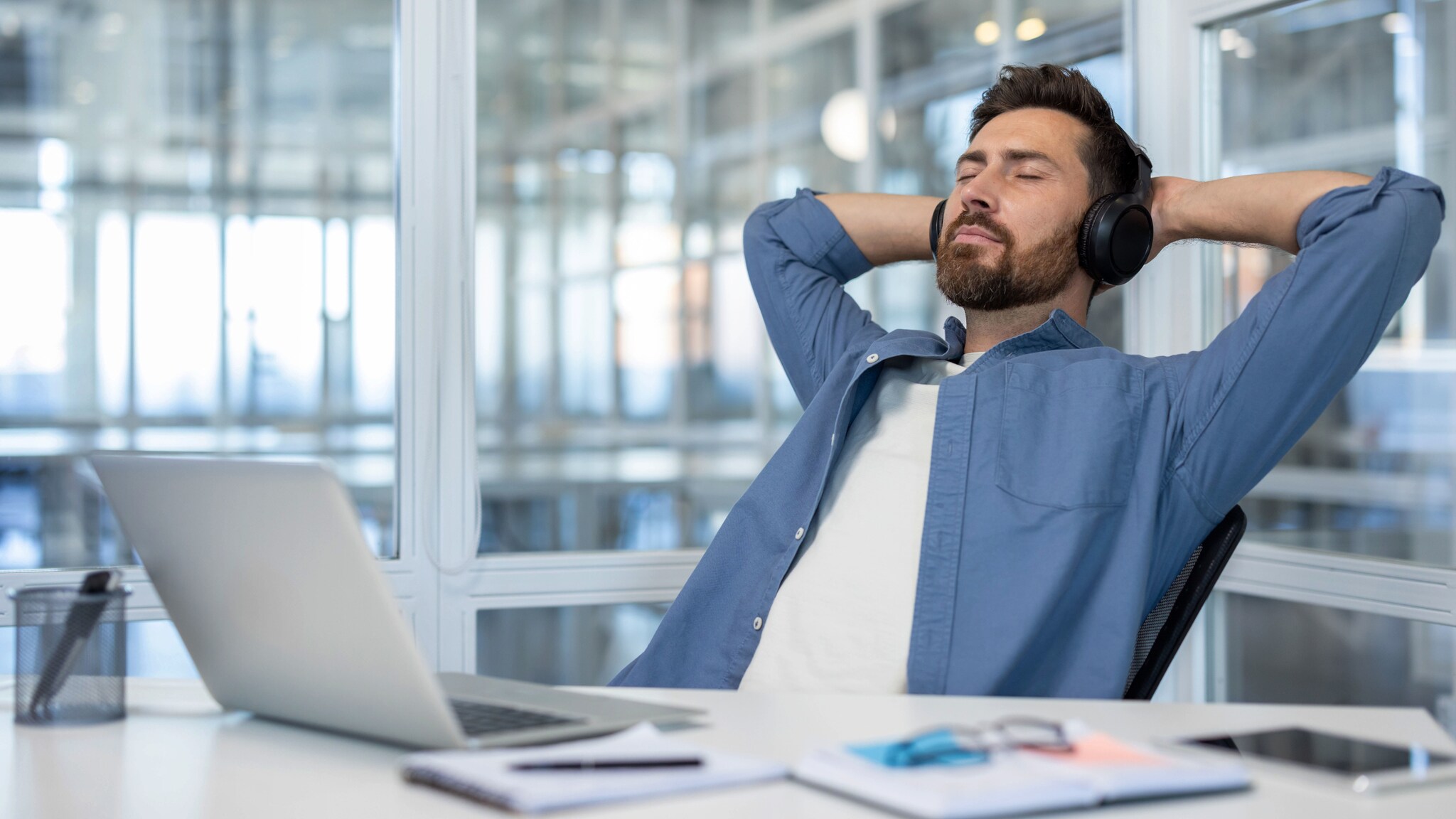Ein Mann im blauen Hemd und mit Kopfhörern lehnt mit geschlossenen Augen und hinter seinem Kopf verschränkten Armen in einem Stuhl an einem Schreibtisch.