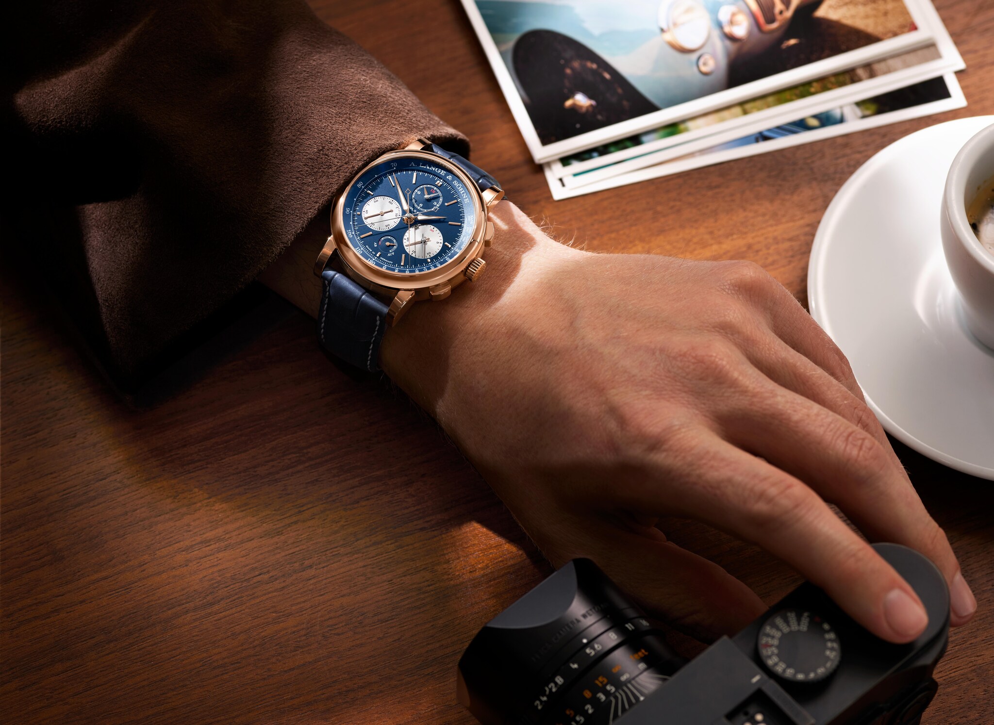 Close-up einer in Blau gehaltenen Uhr von A. Lange und Söhne an einem Männerarm, der auf einem Holztisch mit Fotokamera und Espressotasse liegt Close-up einer in Blau gehaltenen Uhr von A. Lange und Söhne an einem Männerarm, der auf einem Holztisch mit Fotokamera und Espressotasse liegt
