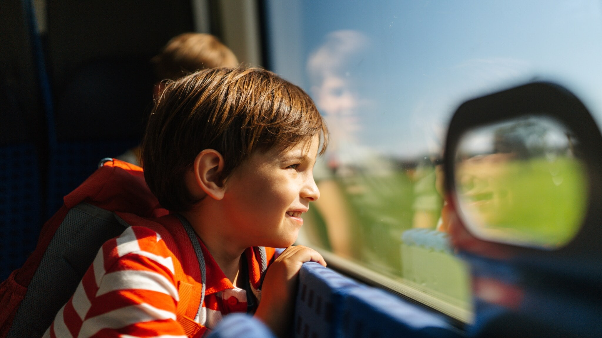 Ein Kind mit gestreiftem Oberteil lehnt sich am Zugfenster und schaut nach draußen. Im Hintergrund ist ein weiteres Kind zu sehen. Ein Kind mit gestreiftem Oberteil lehnt sich am Zugfenster und schaut nach draußen. Im Hintergrund ist ein weiteres Kind zu sehen.