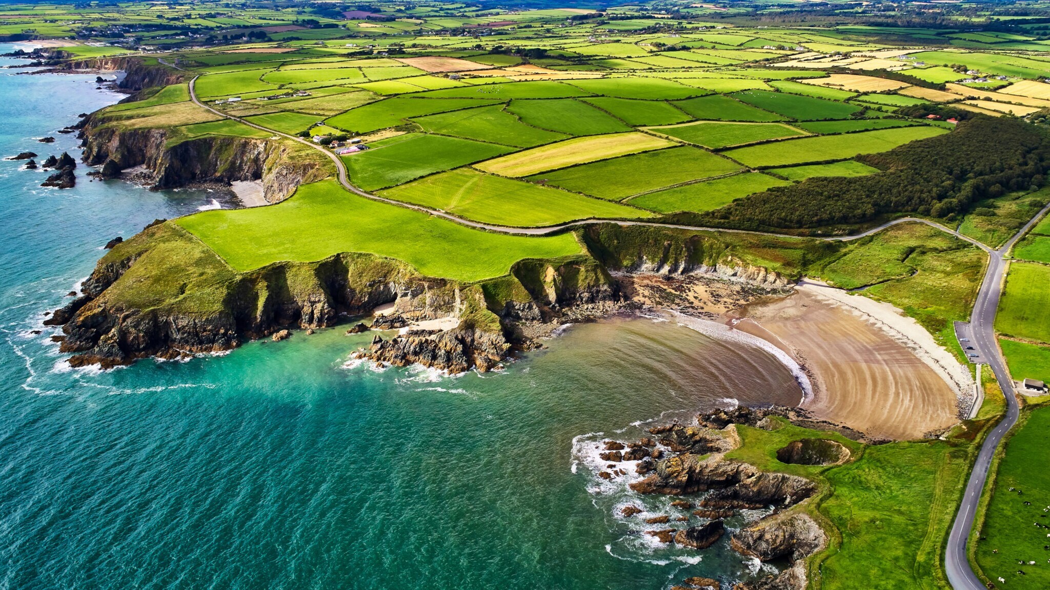 Luftaufnahme der grünen Felder und Küstenklippen an der Copper Coast in County Waterford, Irland, mit Straßen und Bergen im Hintergrund.