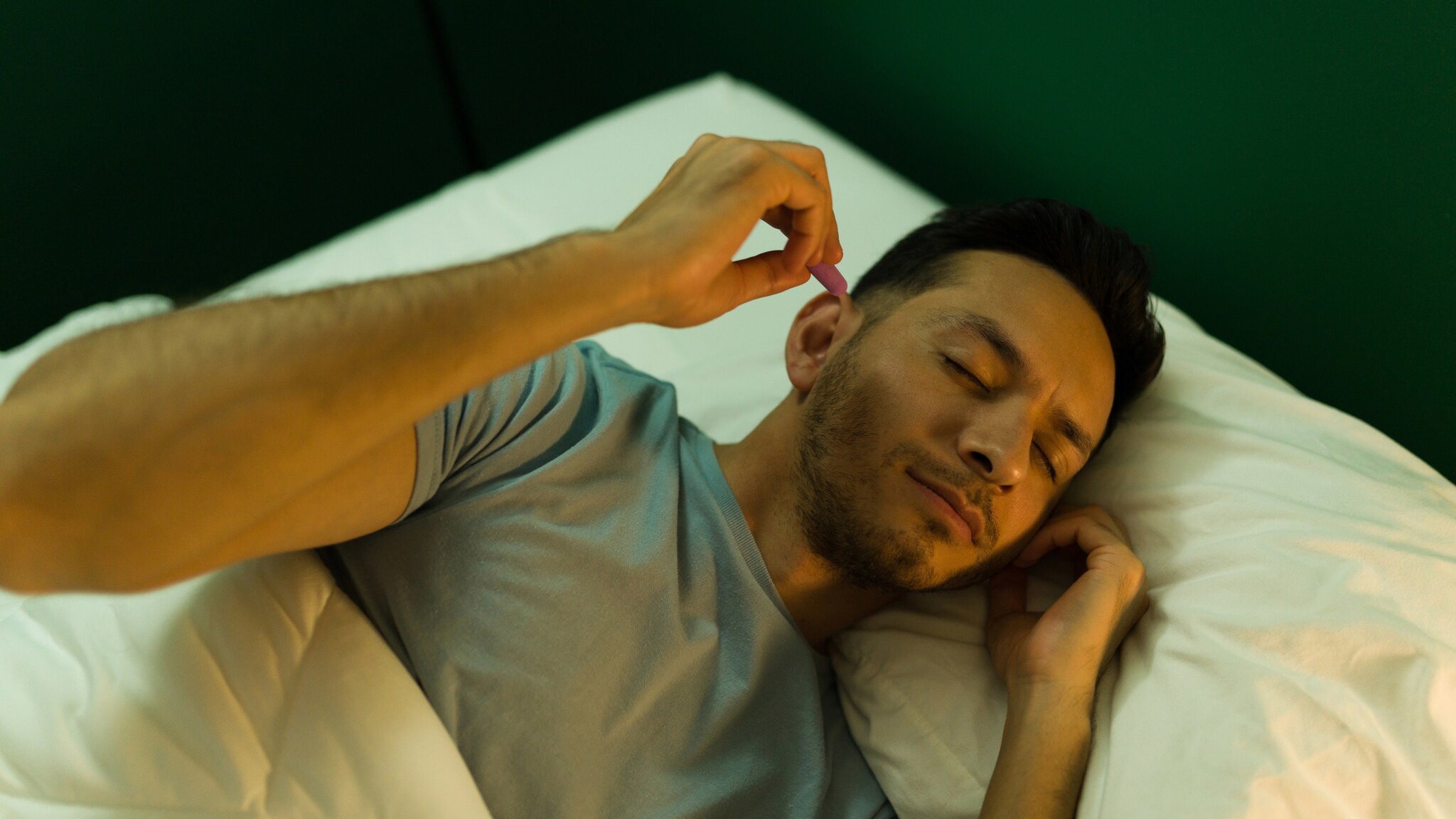 Ein Mann liegt im Bett unter einer weißen Decke, trägt ein graues T-Shirt und steckt sich Ohrenstöpsel ins Ohr. Ein Mann liegt im Bett unter einer weißen Decke, trägt ein graues T-Shirt und steckt sich Ohrenstöpsel ins Ohr.