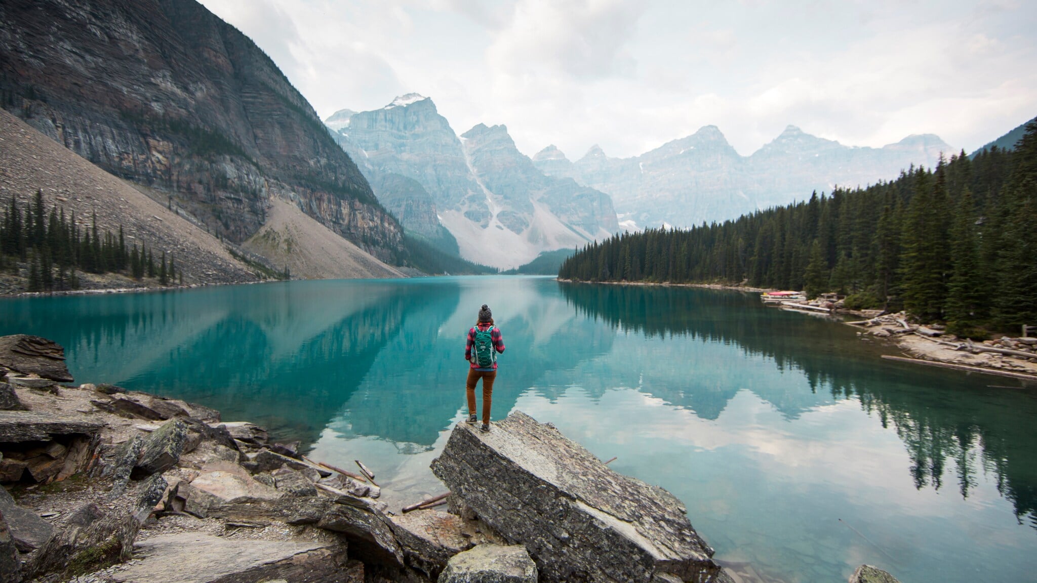 Person mit rotem Rucksack steht auf Felsen am Ufer eines klaren Bergsees, umgeben von Nadelwald und hohen Bergen. Person mit rotem Rucksack steht auf Felsen am Ufer eines klaren Bergsees, umgeben von Nadelwald und hohen Bergen.